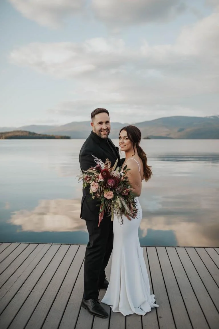 A bride and groom stand on a wooden dock by a calm lake, with mountains and cloudy sky in the background, smiling and holding a large bouquet of flowers.