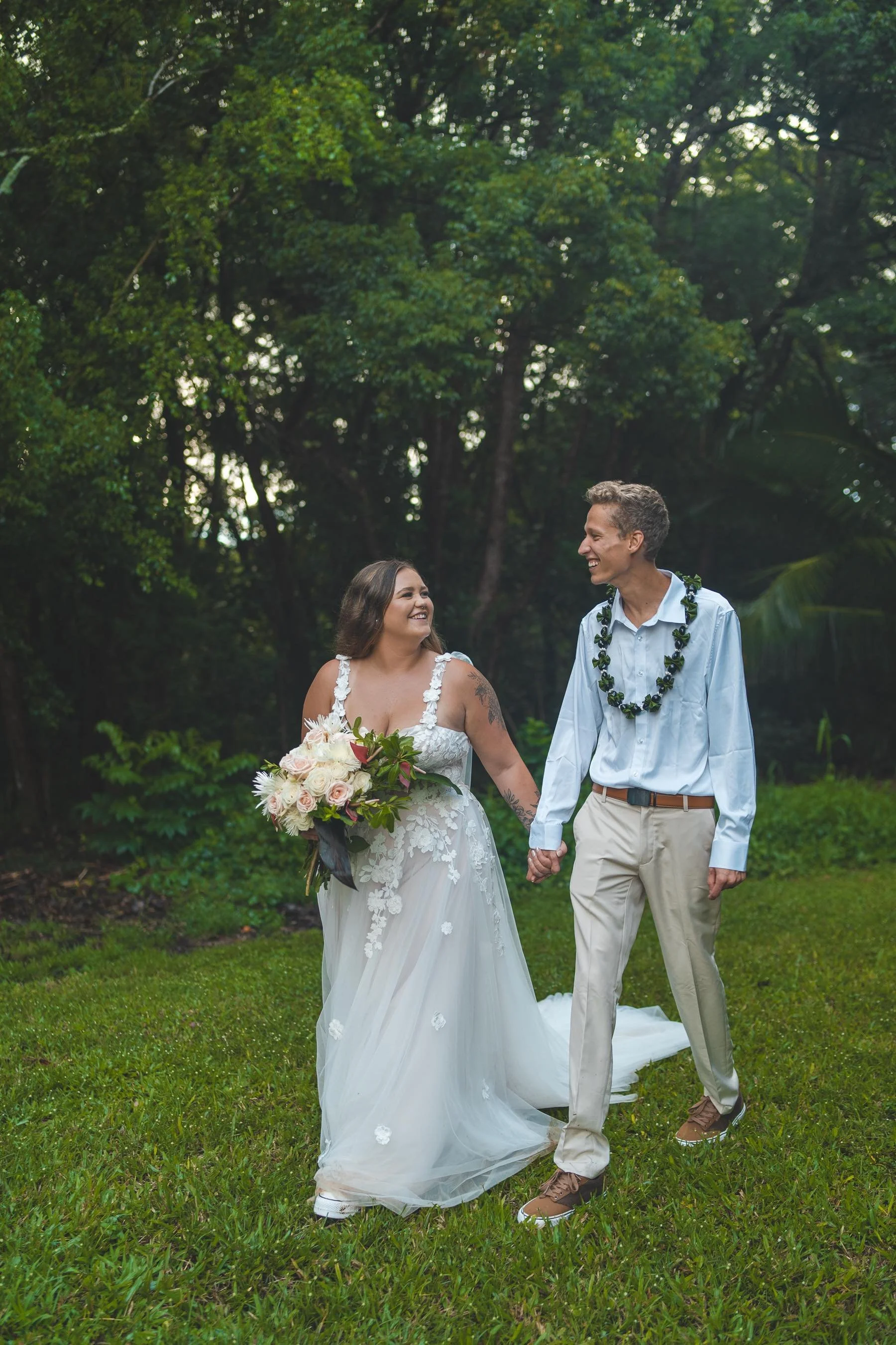 A newlywed couple walking hand in hand on a grassy area, smiling at each other, with lush green trees in the background. The bride is wearing a white wedding dress and holding a bouquet of flowers, and the groom is dressed in a light blue shirt, beige pants, and a green lei.
