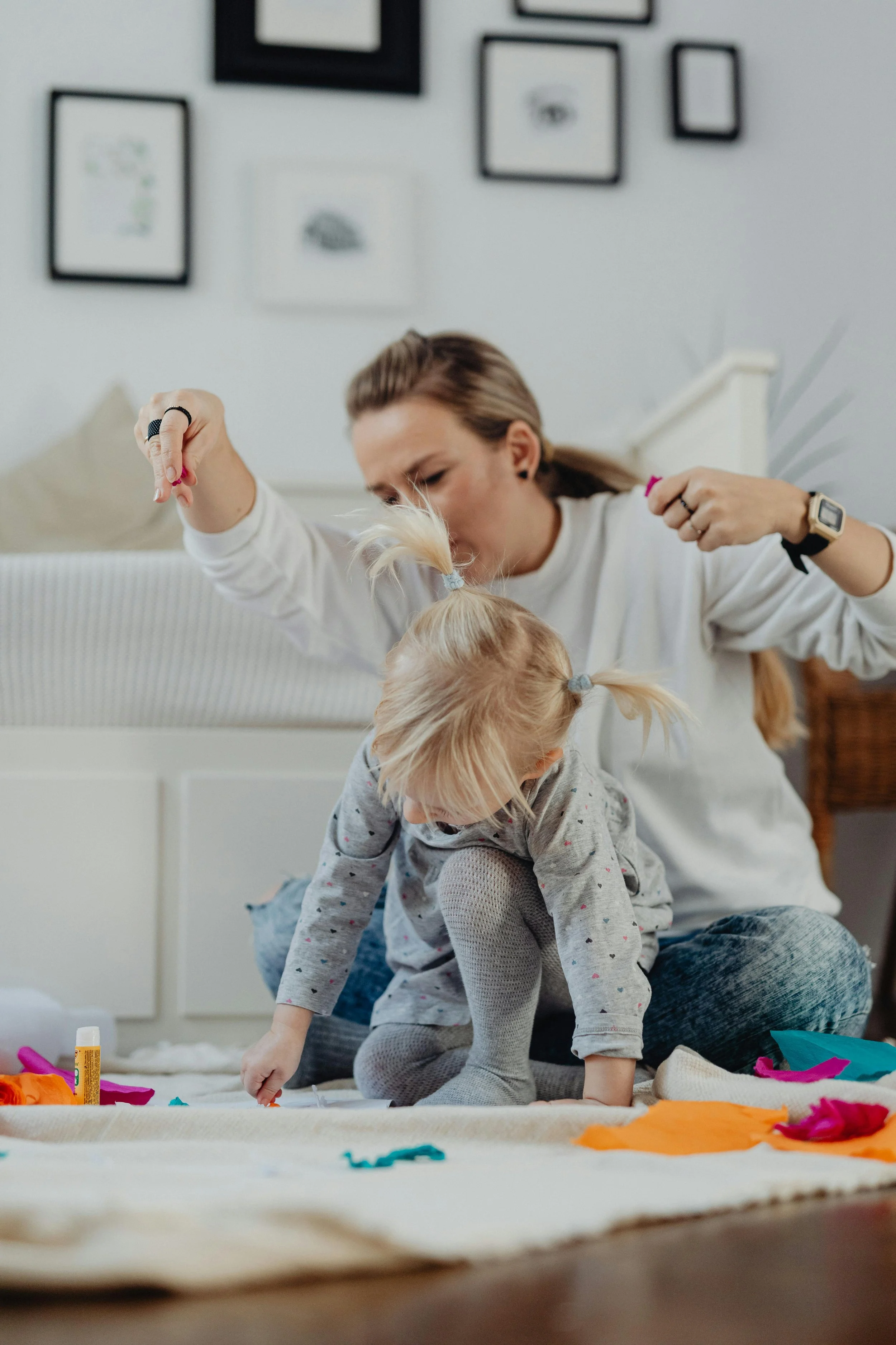 A woman playing with a young girl on the floor, surrounded by colorful fabric, glue, and craft supplies in a cozy room.