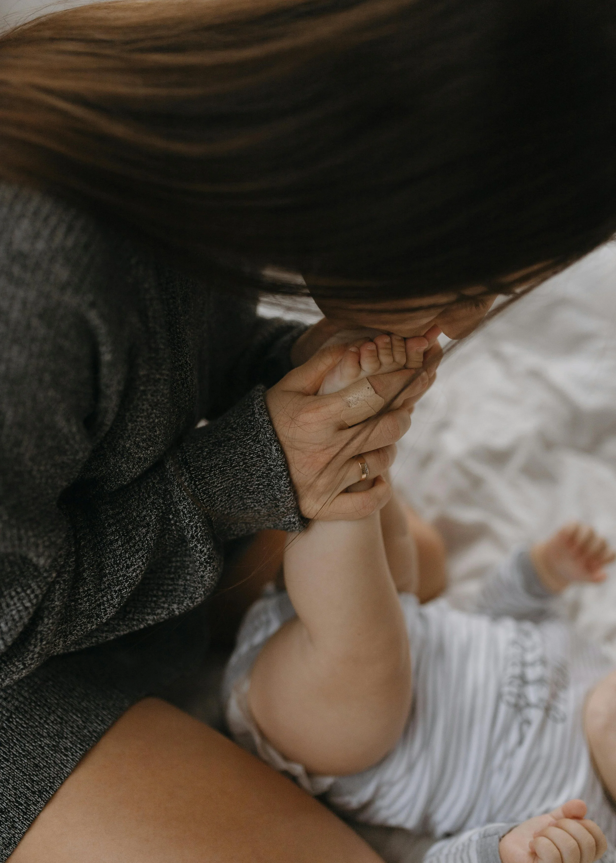 A close-up of a woman with long brown hair holding a baby's small hand, who is lying on her lap wearing a striped onesie, with a white blanket in the background.