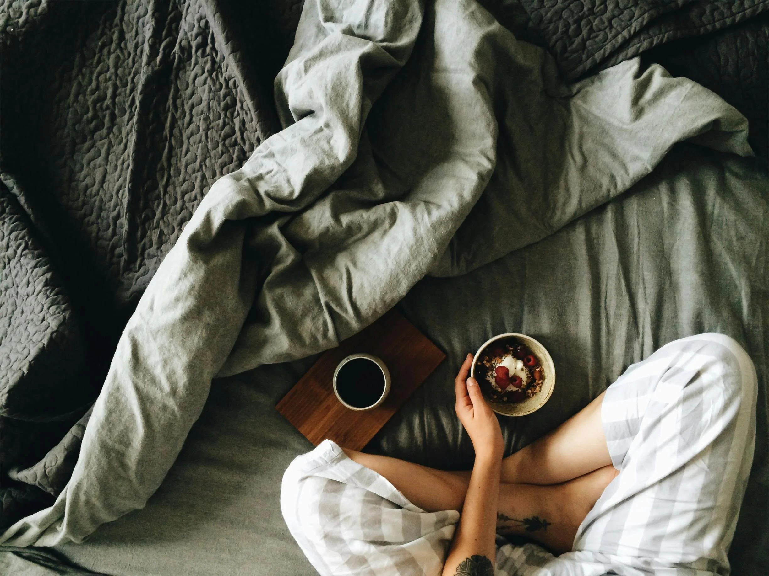 Person holding a bowl of yogurt with berries and nuts, sitting on a bed with gray sheets, next to a cup of coffee and a small wooden tray.