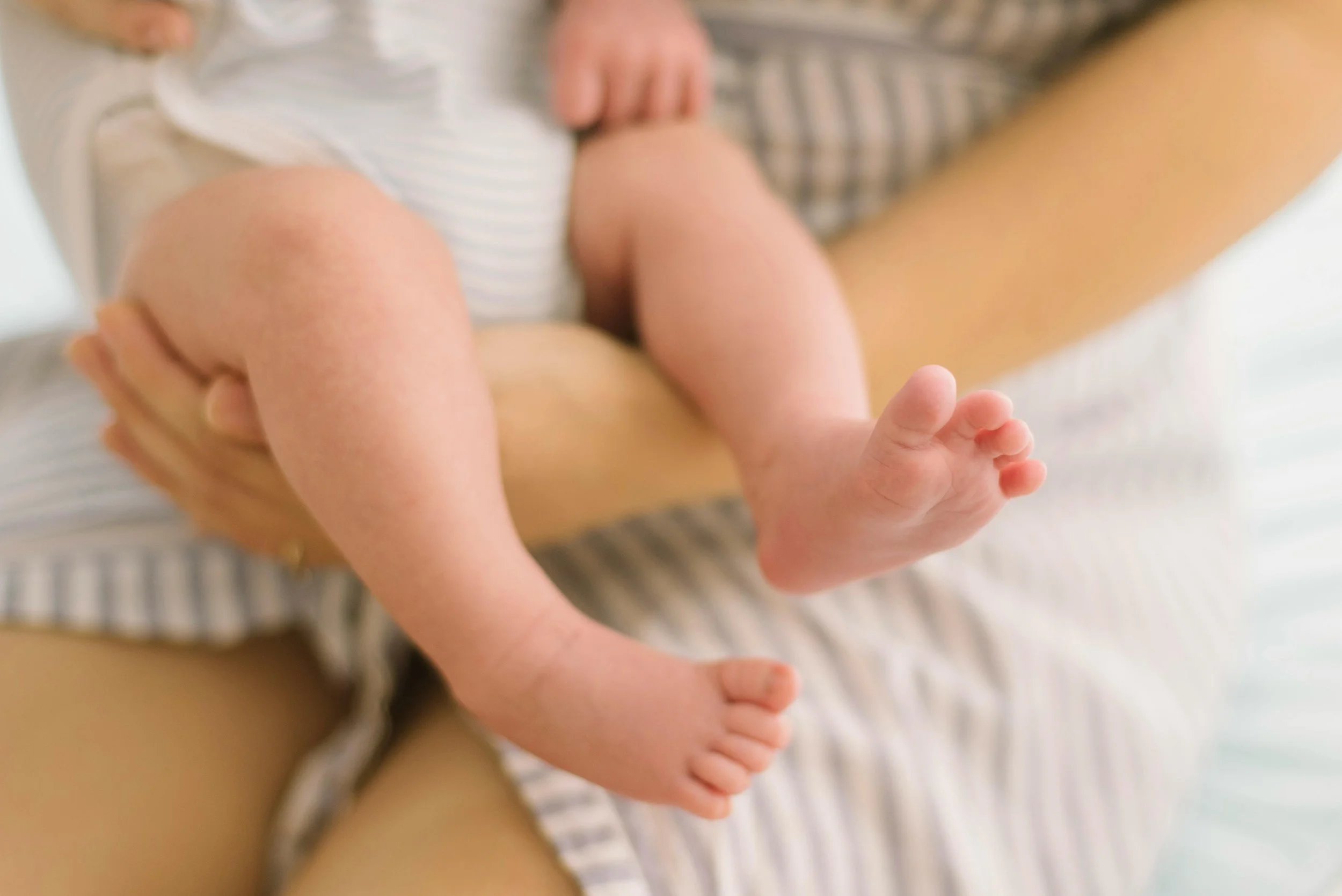 A baby lying on an adult's lap, showing only the baby's legs and feet. The adult is holding the baby with one hand while the baby is dressed in a striped outfit.