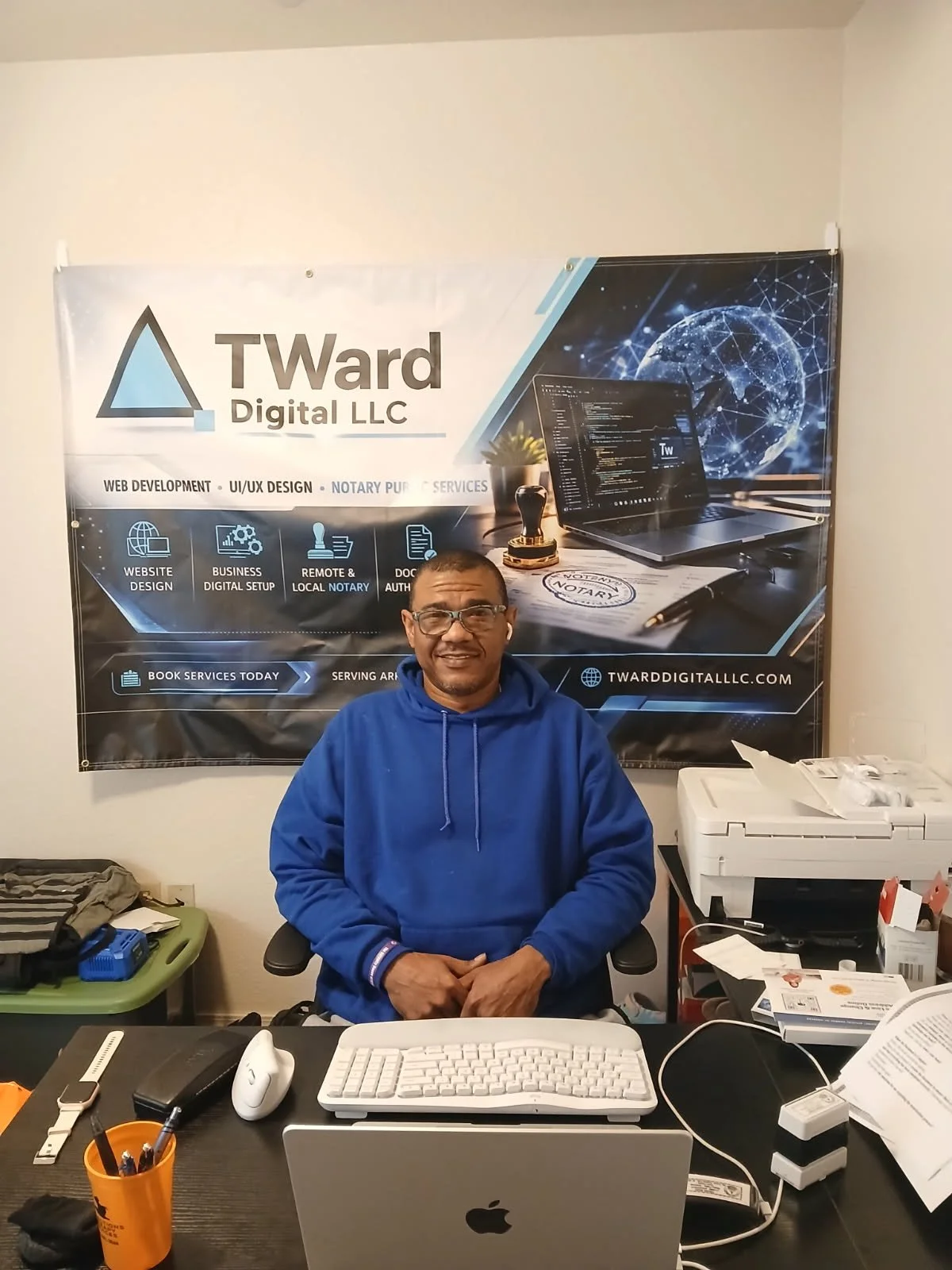 A man sitting at a cluttered office desk, with a computer monitor, keyboard, mouse, and various office supplies, in front of a banner for Tward Digital LLC.