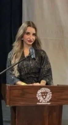 Joven mujer en escenario, de cabello largo y rubio, con vestido oscuro, de pie detrás de un podio de madera con un escudo de león.