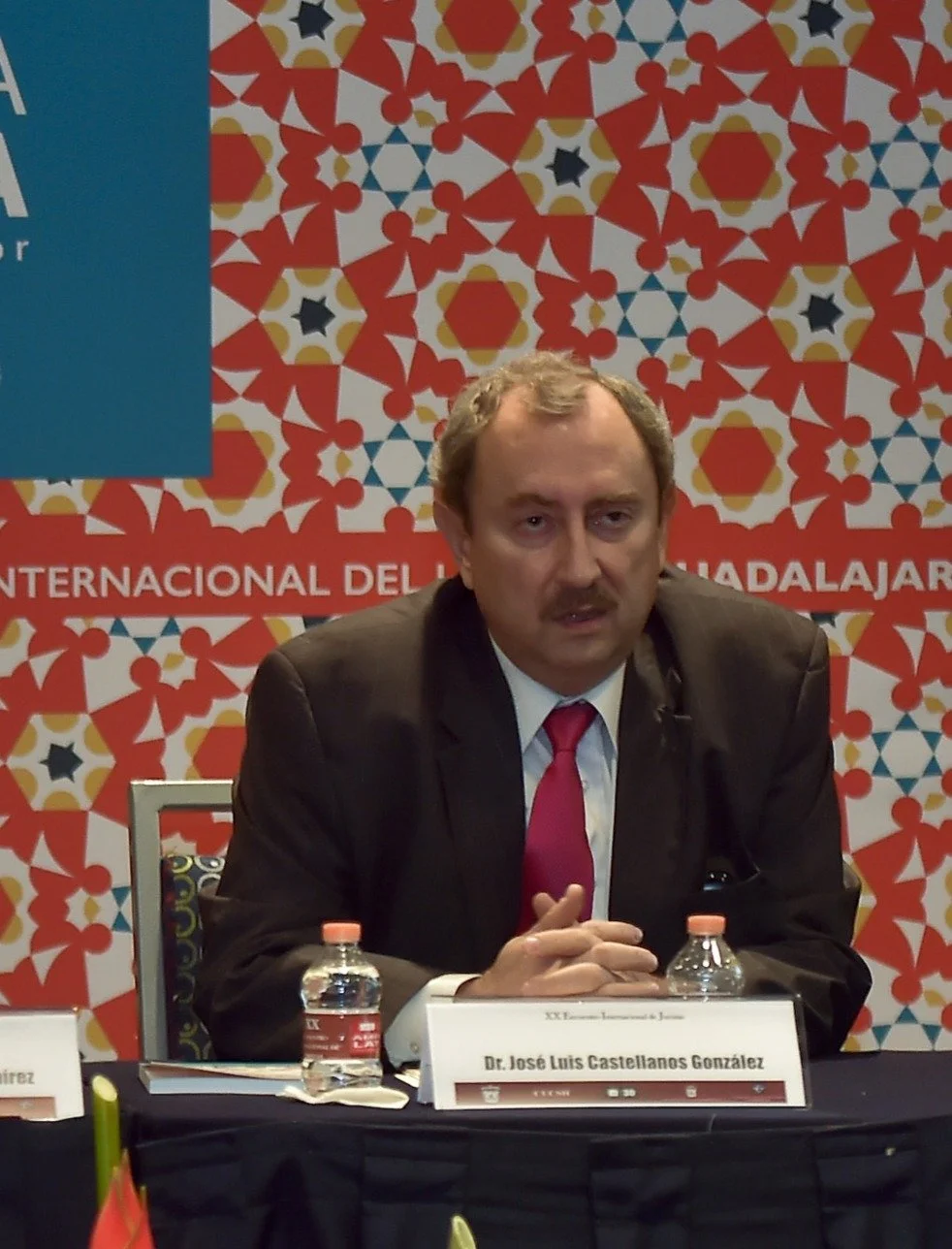 Hombre de traje y corbata sentado en una mesa con fondo de patrón colorido y cartel con texto en español, probablemente en un evento o conferencia.