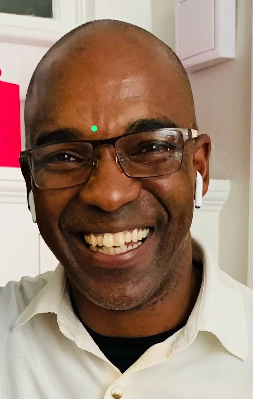 A smiling man with glasses, wireless earbuds, a semi-shaved head, wearing a white shirt, indoors.