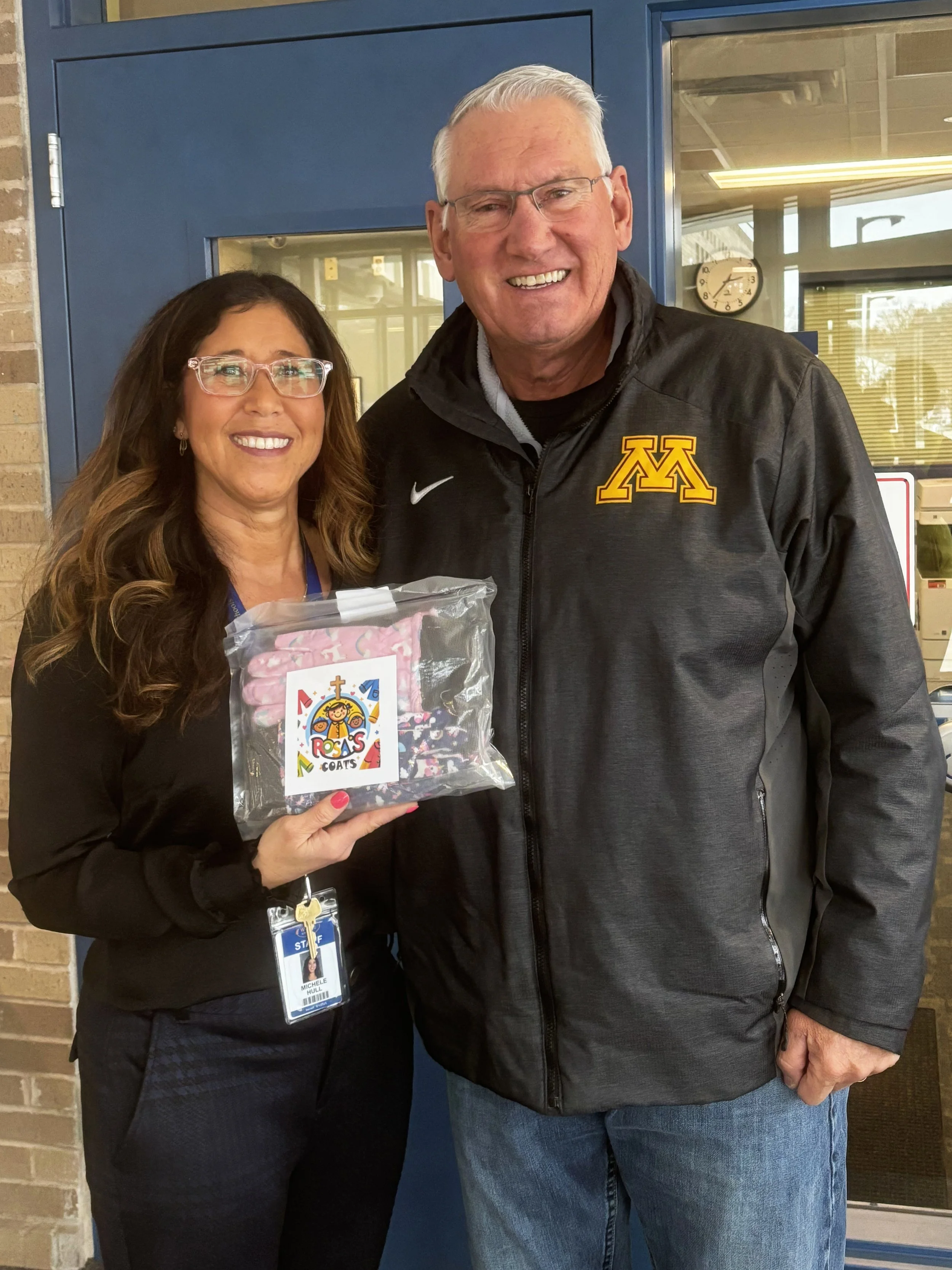 A woman and a man smiling and posing together inside a building. The woman is holding a clear plastic bag with a colorful label that says 'Rosa's Coats.' The woman is wearing glasses and an ID badge, and the man is wearing a black jacket with a University of Minnesota logo.