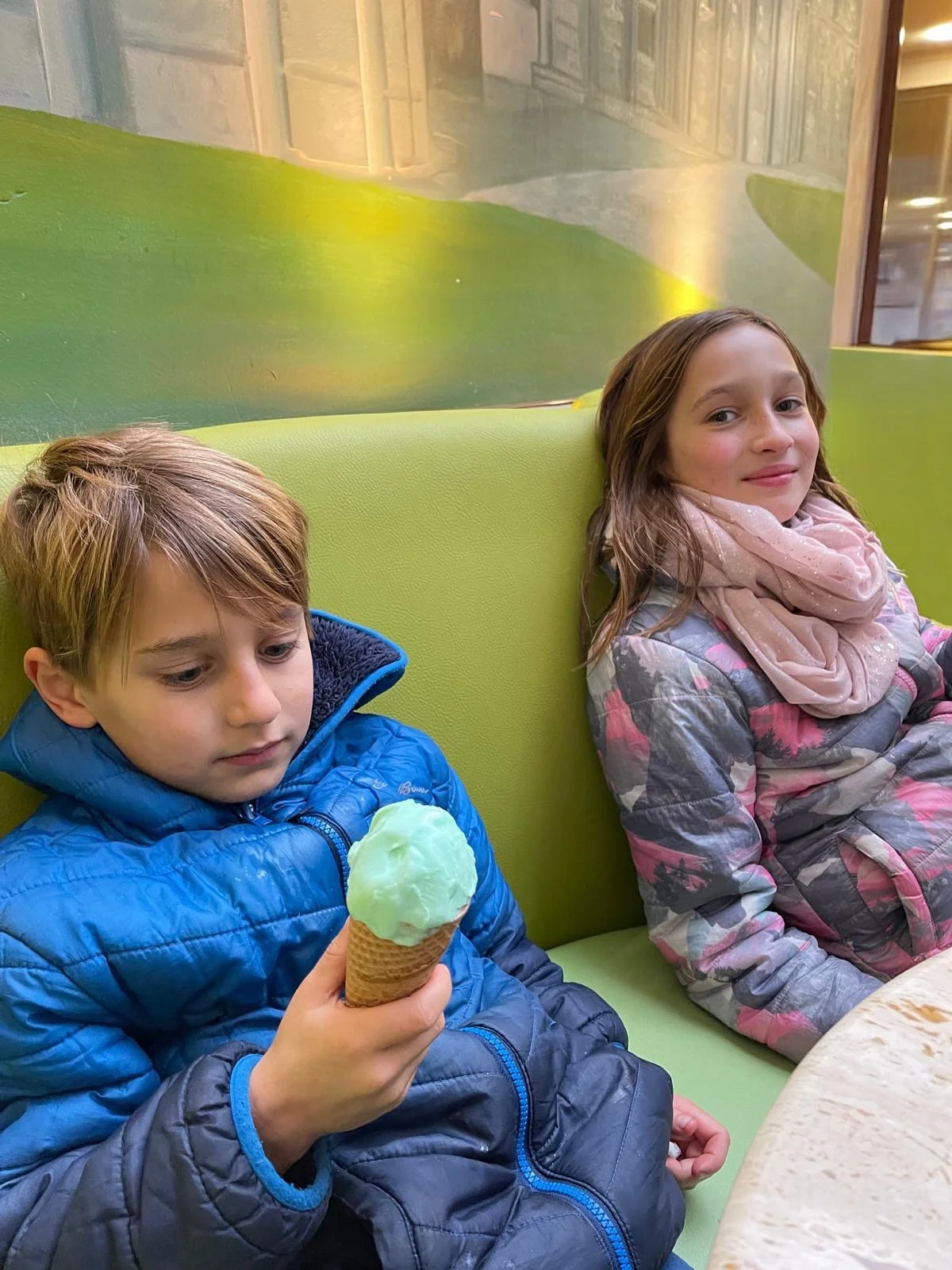 A young boy in a blue jacket holding a green ice cream cone, sitting next to a girl in a pink and gray jacket with a pink scarf, both sitting on a lime green bench inside a building.