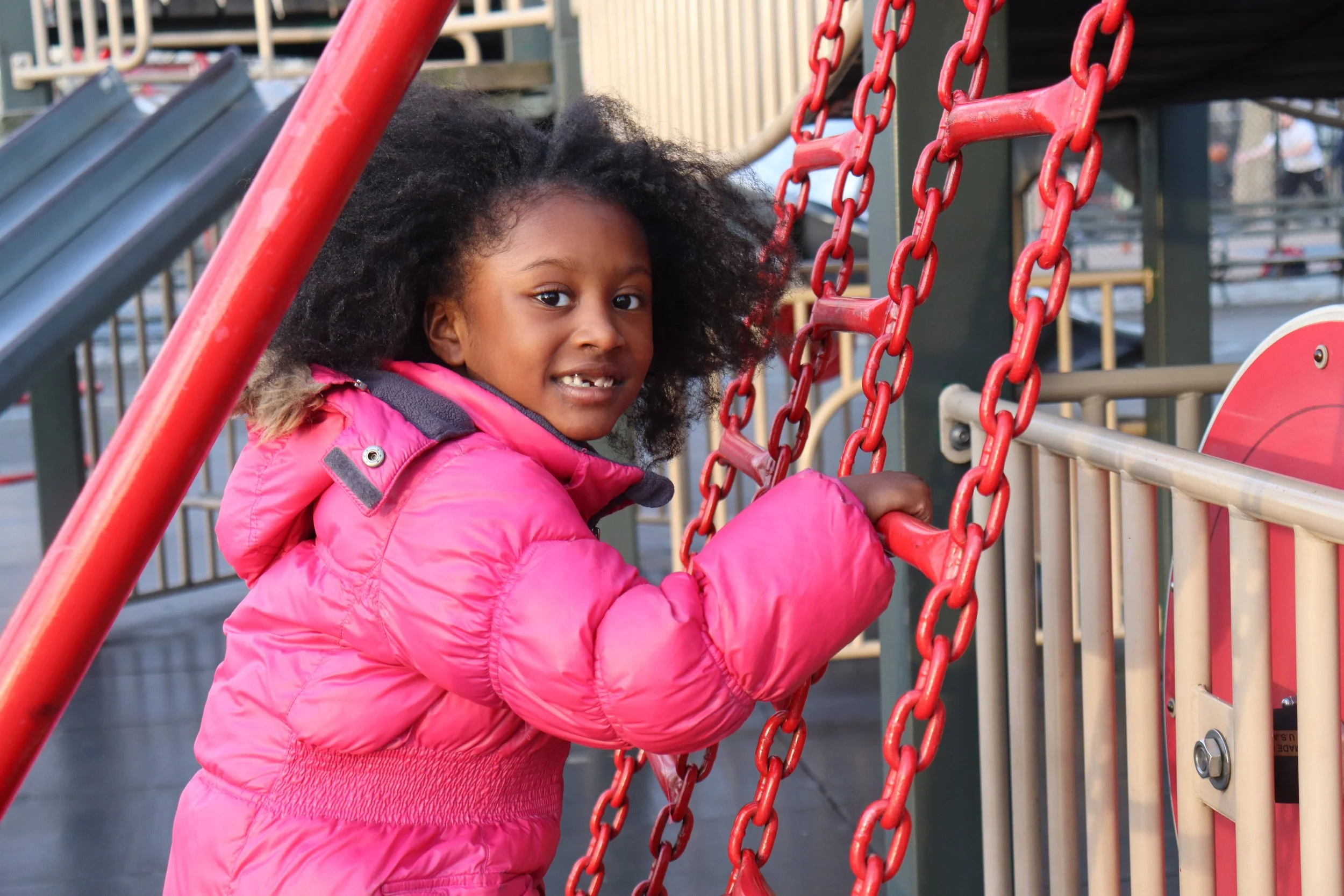 Girl in a pink jacket holding onto red chains on a playground.