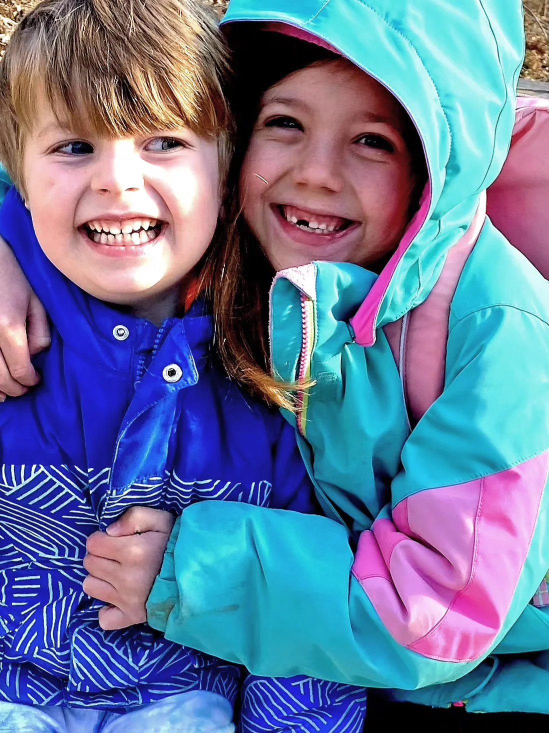 Two young girls smiling and hugging outdoors, dressed in colorful winter jackets, with a natural background.