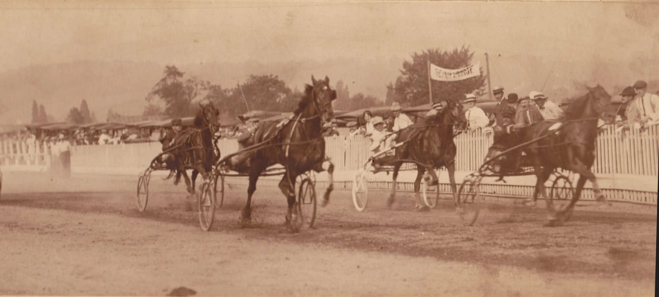 Sepia photograph of horses and riders racing on a dirt track, with spectators lining the fence near Ridgewood, NJ