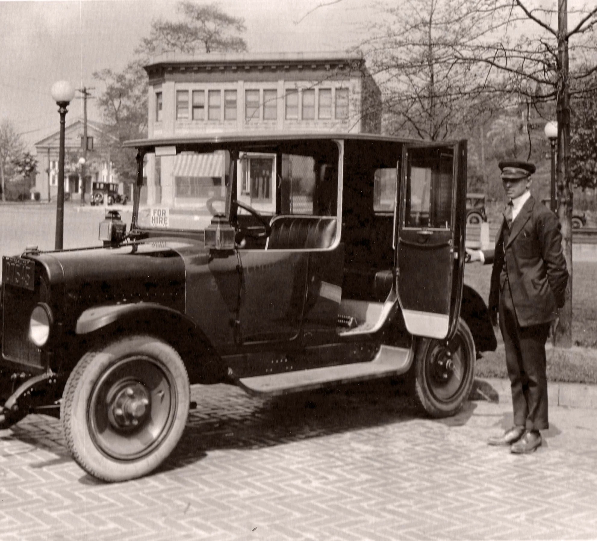 Black and white photograph of a uniformed driver standing beside a vintage for-hire automobile on a cobblestone street, Ridgewood, NJ