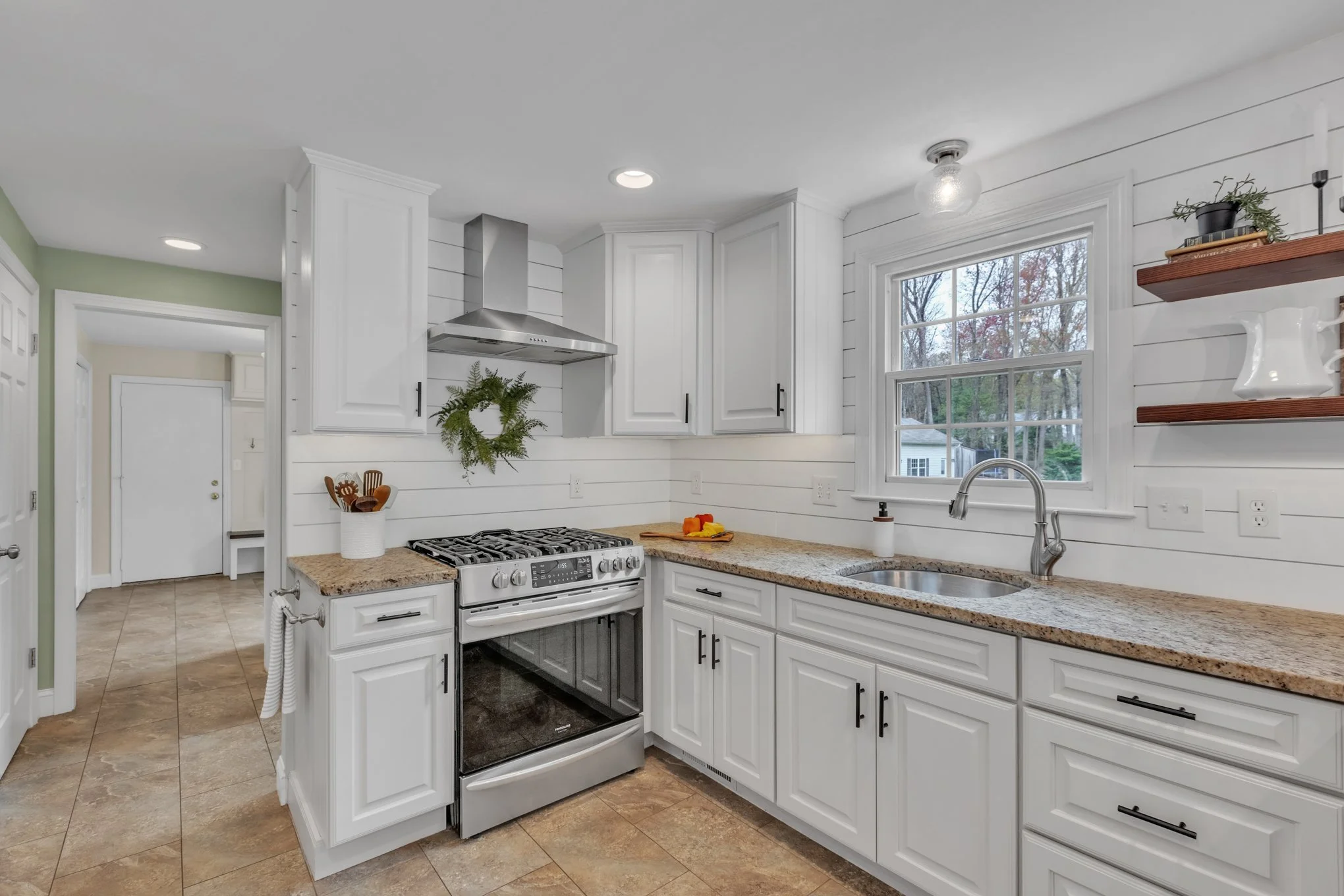 A modern kitchen with white cabinets, granite countertops, and stainless steel appliances. Features include a stove with a range hood, a window above the sink, and floating shelves with decorative items.