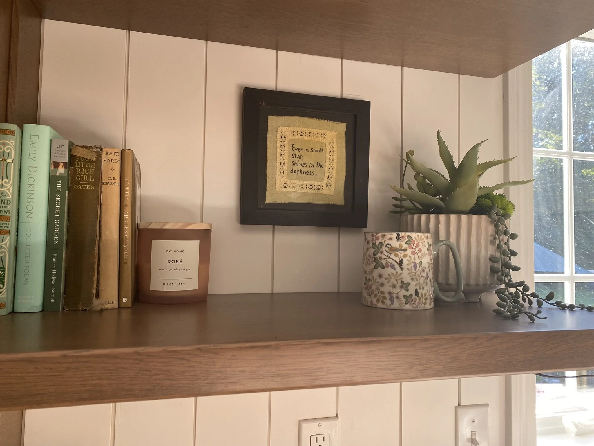 A wooden shelf decorated with books, a small candle, framed embroidery, and potted succulents near a window with sunlight.