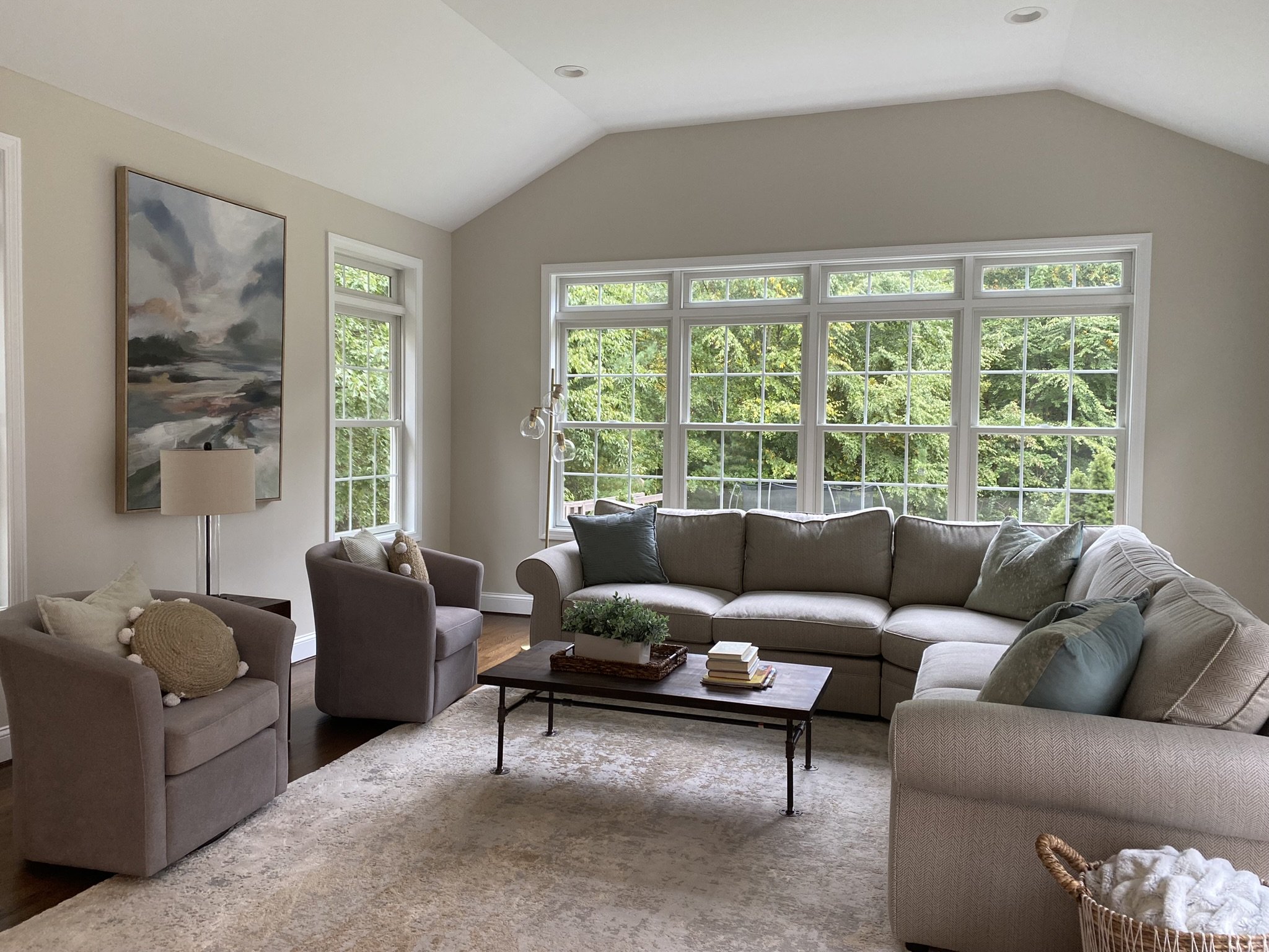 Living room with large beige sectional sofa, two armchairs, a coffee table with books and a plant, large windows with greenery outside, abstract painting on wall, beige walls, and a plush rug.