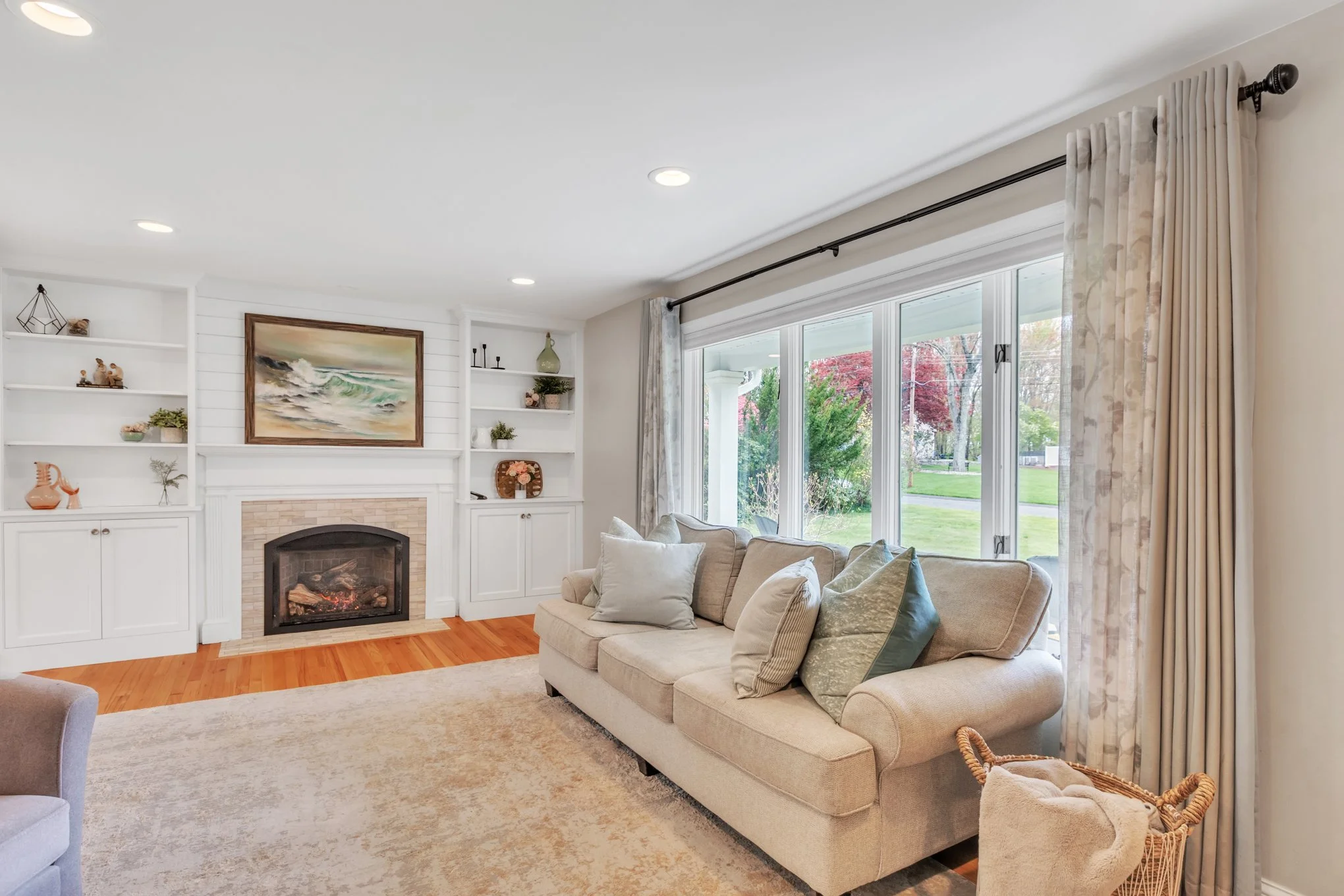 Living room with a beige sofa, multiple pillows, large window with curtains, built-in white shelves, a fireplace, and hardwood flooring.