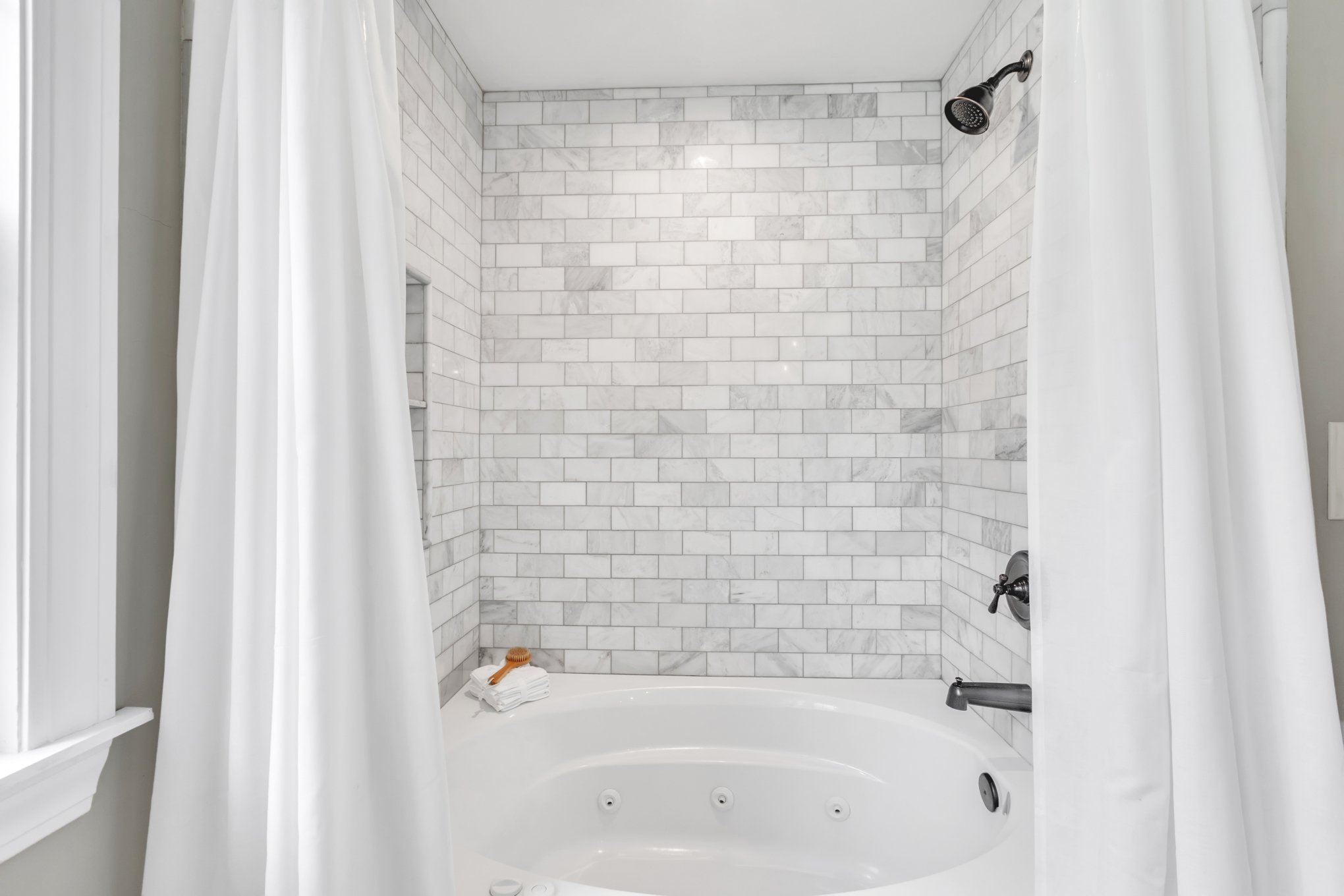 White bathtub with jets, surrounded by white marble tile walls, curtains on both sides, with a stack of towels and a brush on the edge.