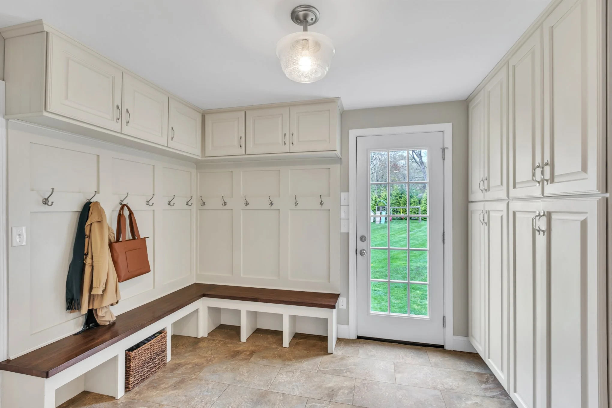 A spacious mudroom with built-in white cabinets, hooks for coats, a wooden bench, and a view of a green backyard through a glass door.