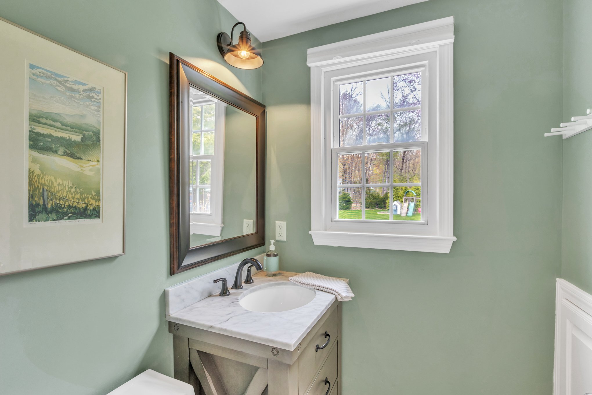 Bathroom with green walls, white window and trim, rectangular mirror above marble vanity, sink faucet, soap dispenser, towel, and a picture of a landscape on the wall.