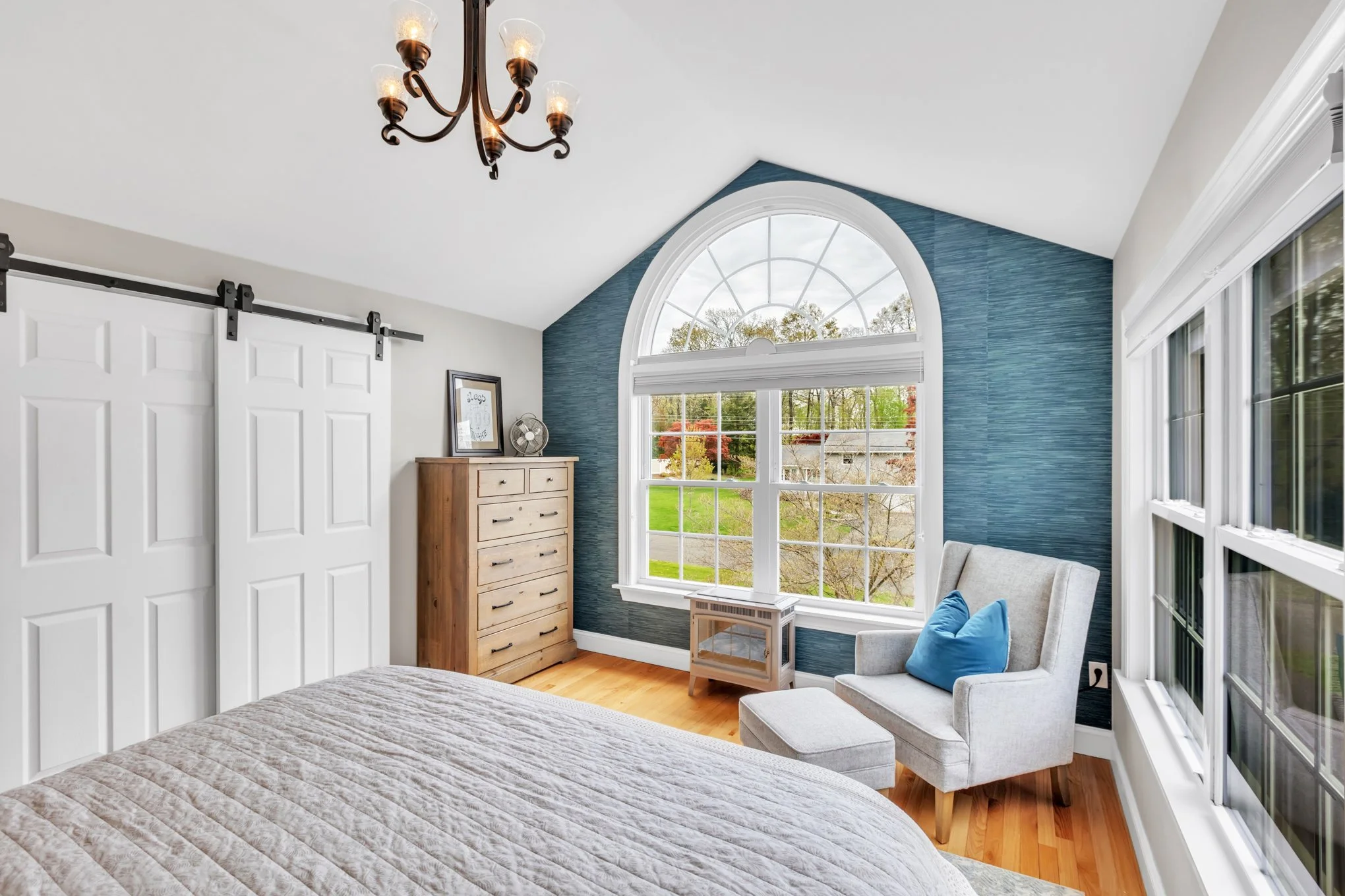 A bedroom with a large arched window showing trees outside, a white armchair with a blue pillow, a wooden dresser, white sliding closet doors, a chandelier, and light-colored hardwood flooring.