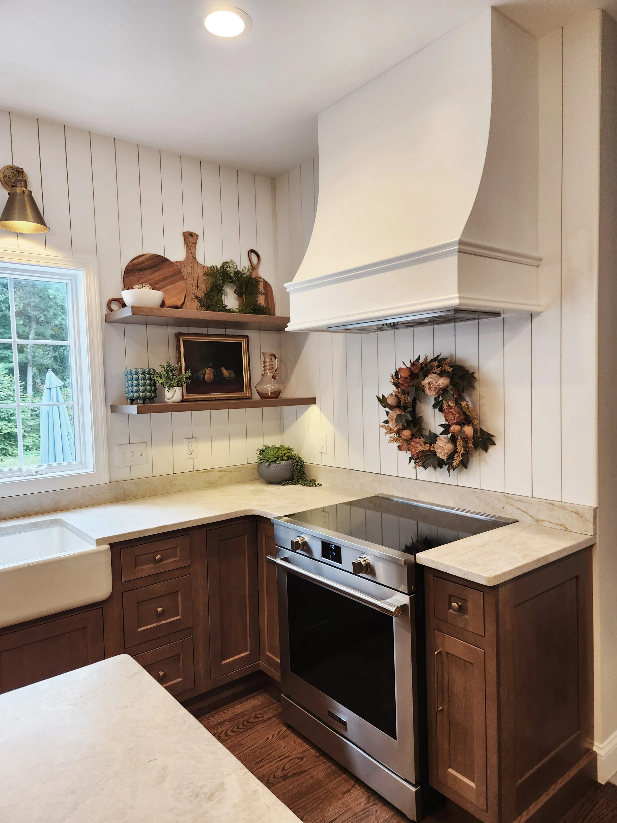 Kitchen corner with wooden cabinets, a beige countertop, a white farmhouse sink, a black stove, a window with outdoor view, and decorative shelves with plants, artwork, and kitchenware.
