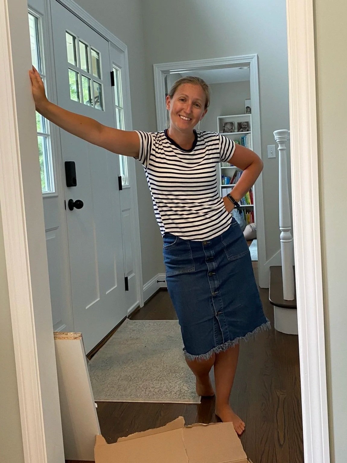 A woman standing in a doorway inside a house, smiling, wearing a striped t-shirt and a denim skirt, with one arm extended and one hand on her hip.