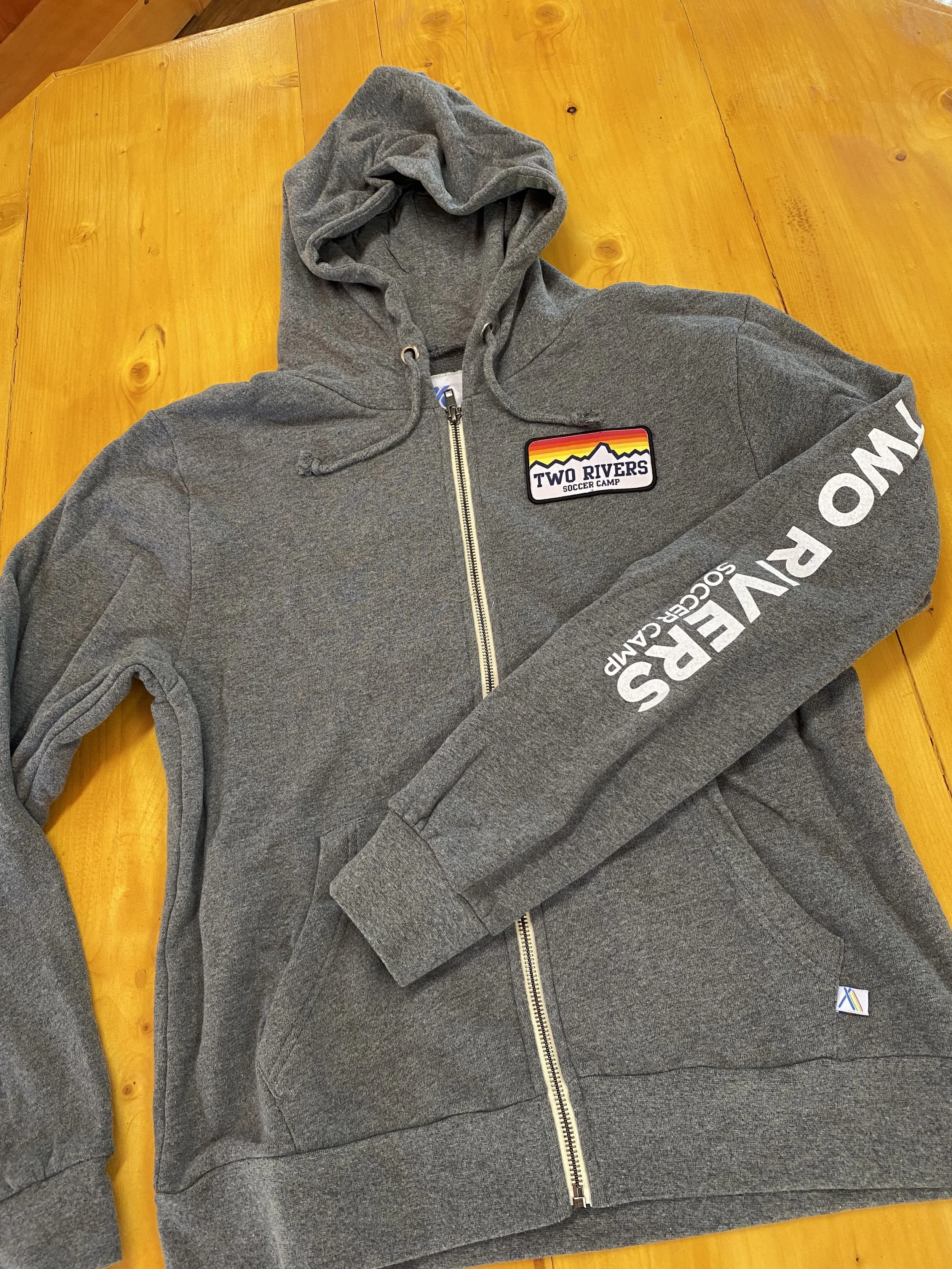 Gray zip-up hoodie with a patch that says "Two Rivers Soccer Camp" and a mountain logo, lying on a wooden surface.