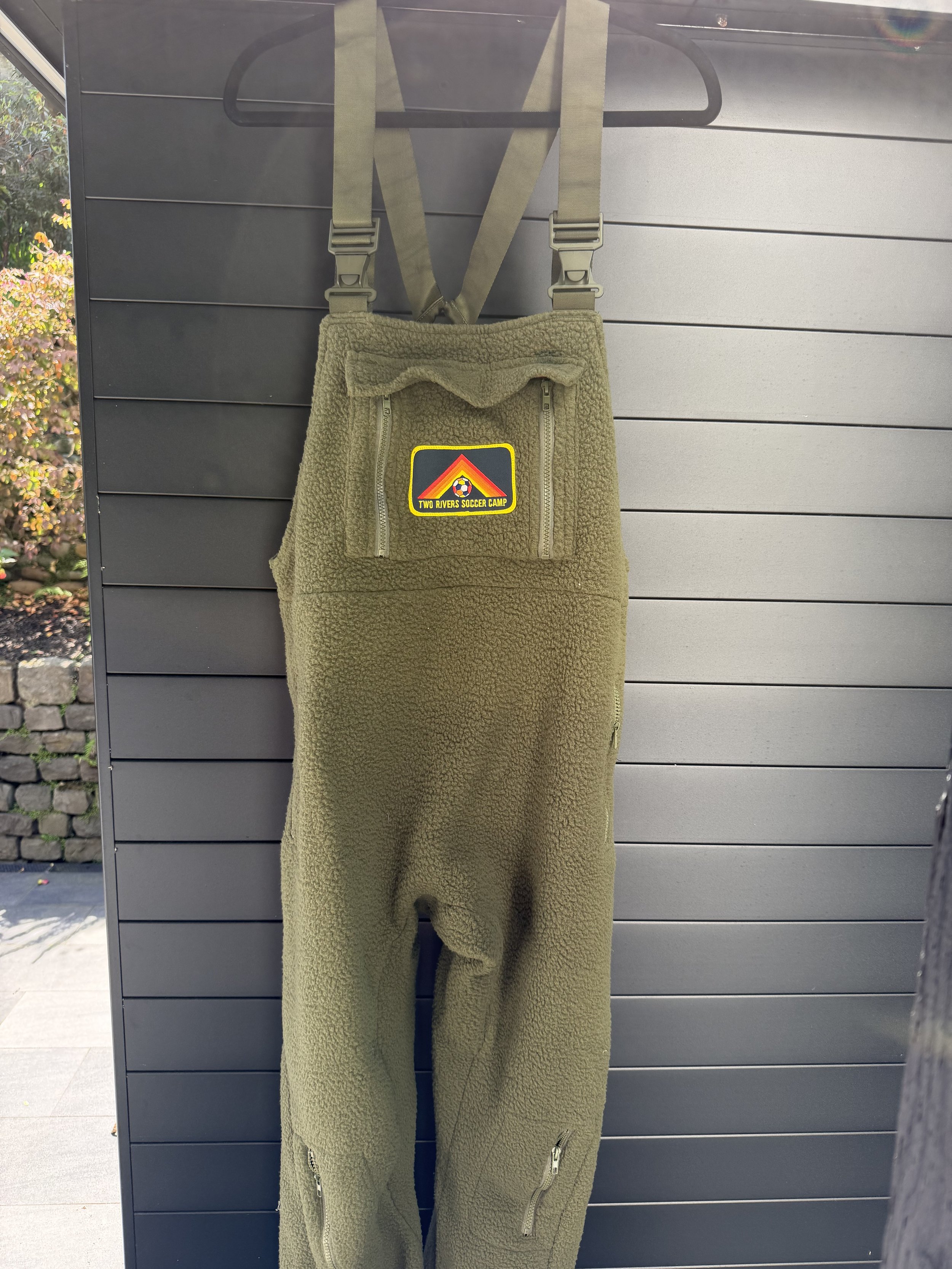 Olive green fleece overalls with zippered pockets hanging on a black hanger against a gray wall, with a patch that says 'Two Rivers Soccer Camp'.