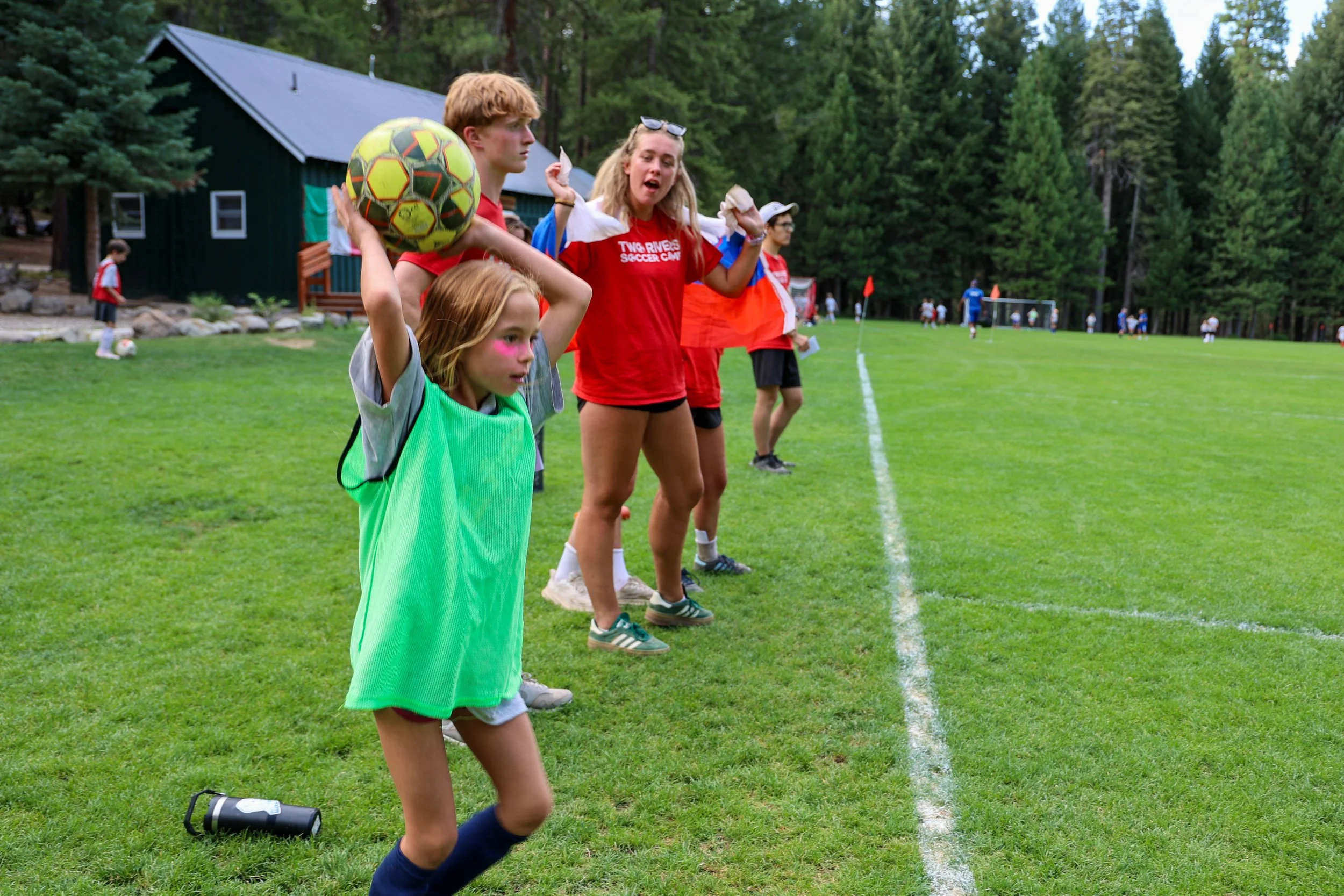 Group of children on the sidelines of a soccer field, some cheering, other waiting with a soccer ball, surrounded by trees.