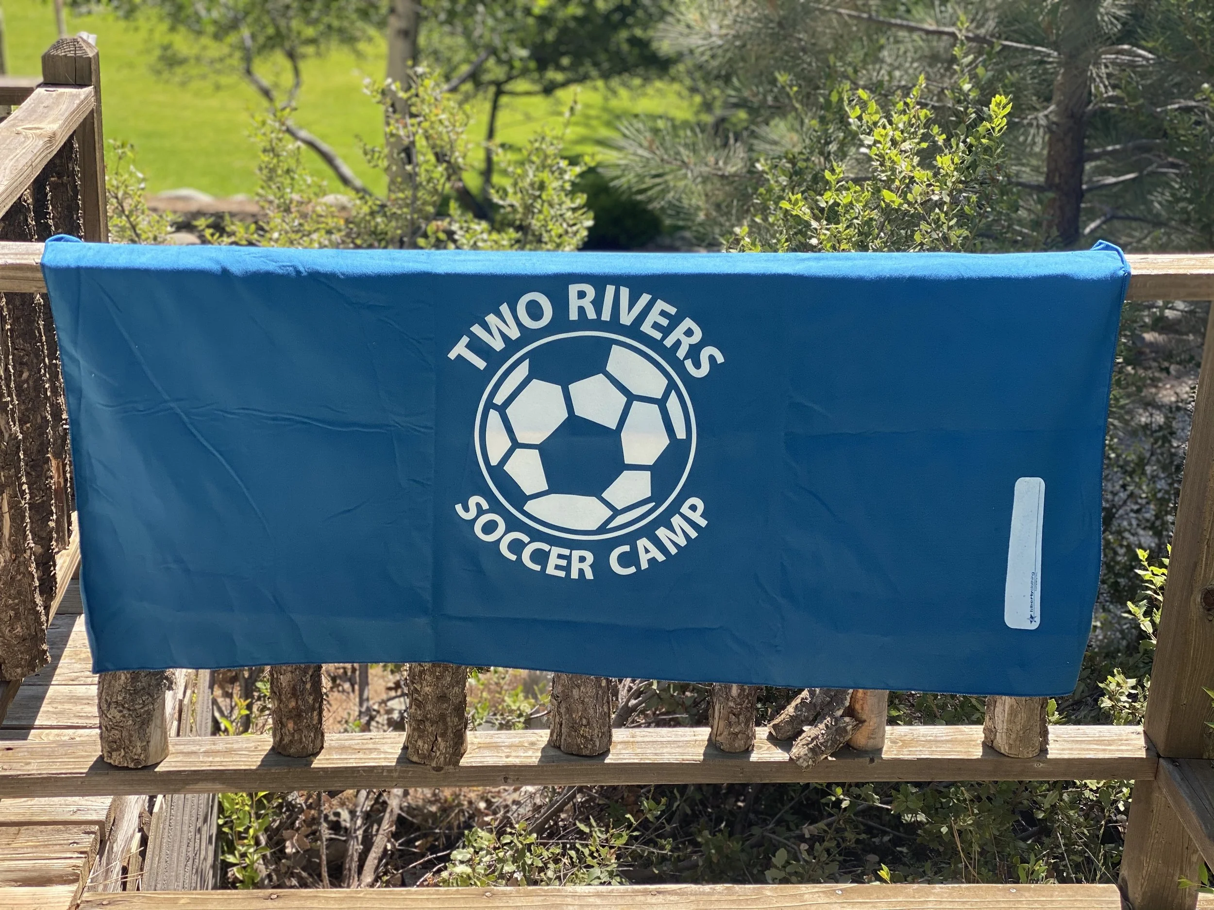 Blue towel with white logo and text that reads 'Two Rivers Soccer Camp,' hanging on a wooden railing outdoors.