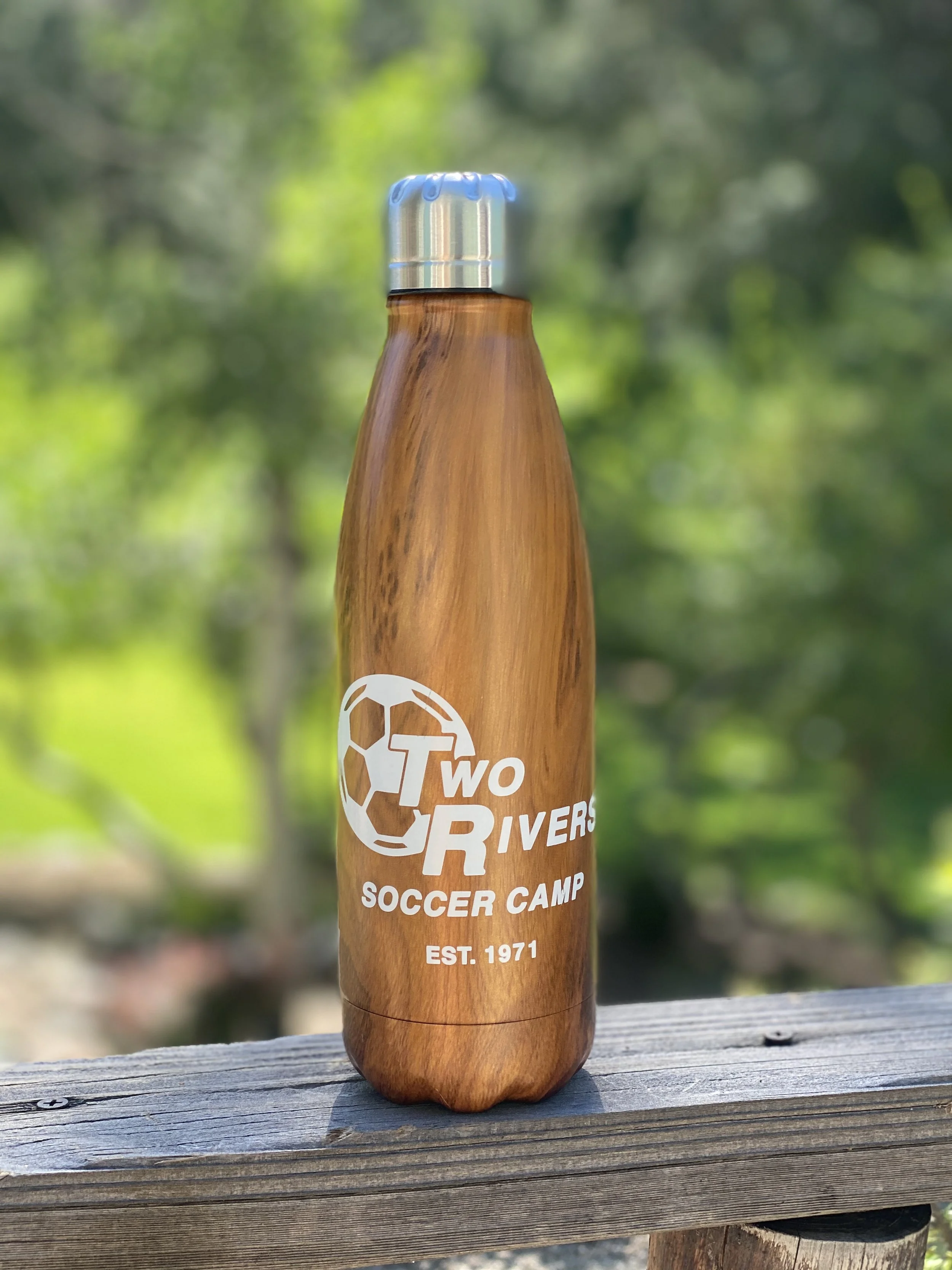 A brown wooden water bottle with a metallic cap, featuring a logo and text for Two Rivers Soccer Camp, established in 1971, placed on a wooden surface outdoors with green blurred trees in the background.