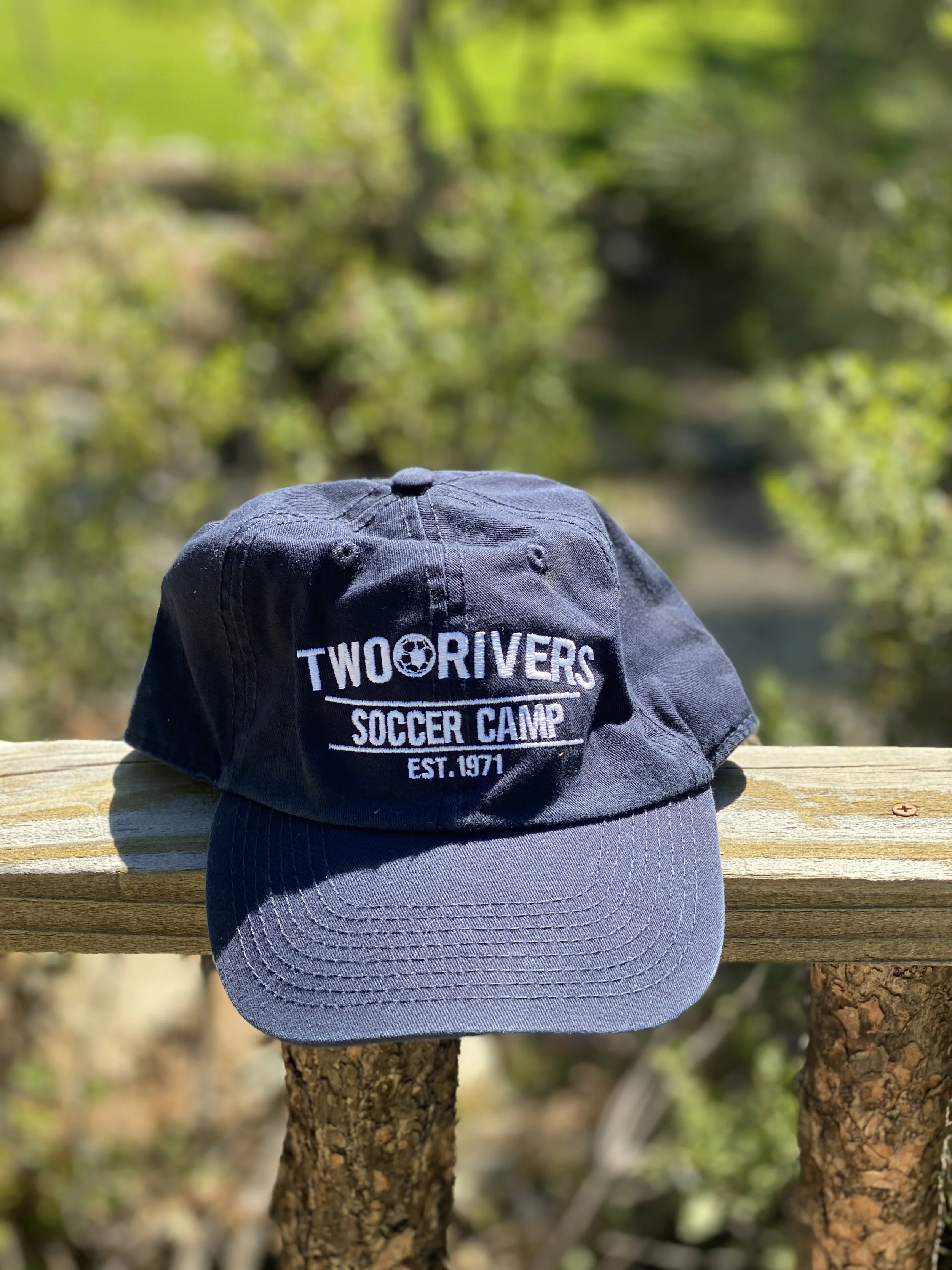 A black cap with white embroidery reading "TWO RIVERS SOCCER CAMP EST.1971" on a wooden railing outdoors with greenery in the background.
