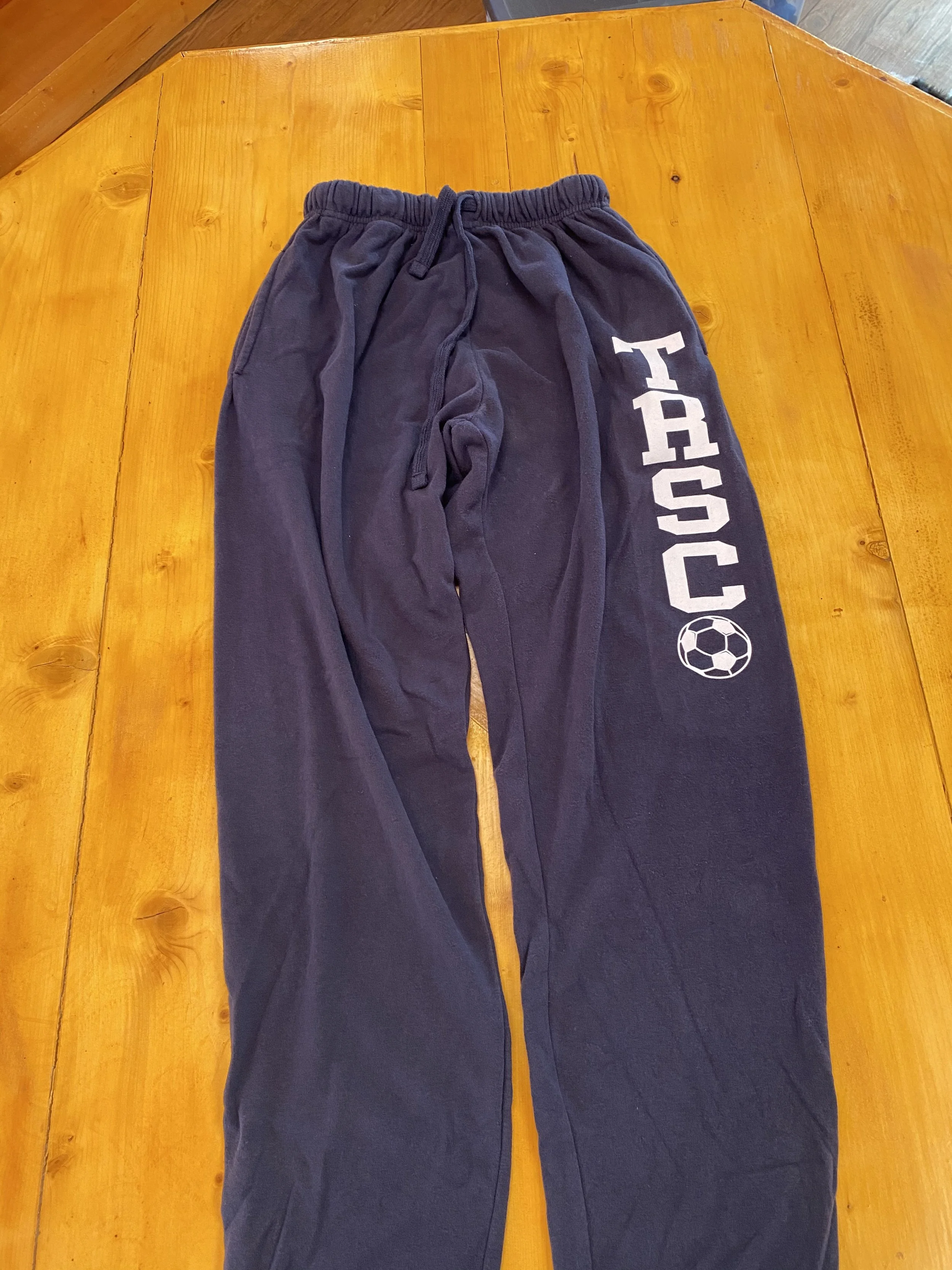 Pair of sweatpants with white text and soccer ball graphic on the right leg, laid flat on a wooden table.