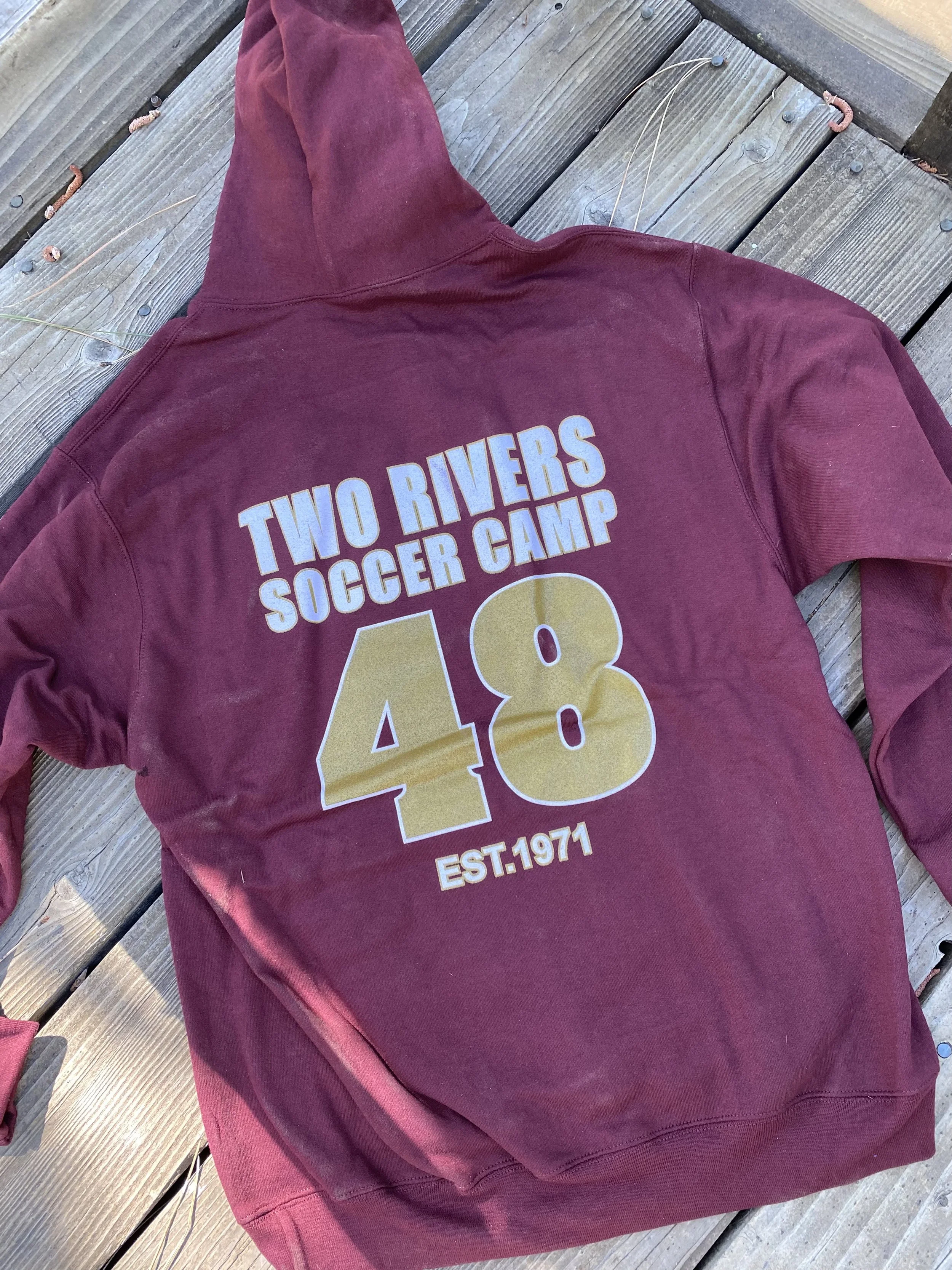 Maroon hoodie with yellow and white text that reads 'Two Rivers Soccer Camp 48 Est.1971' lying on a wooden deck.