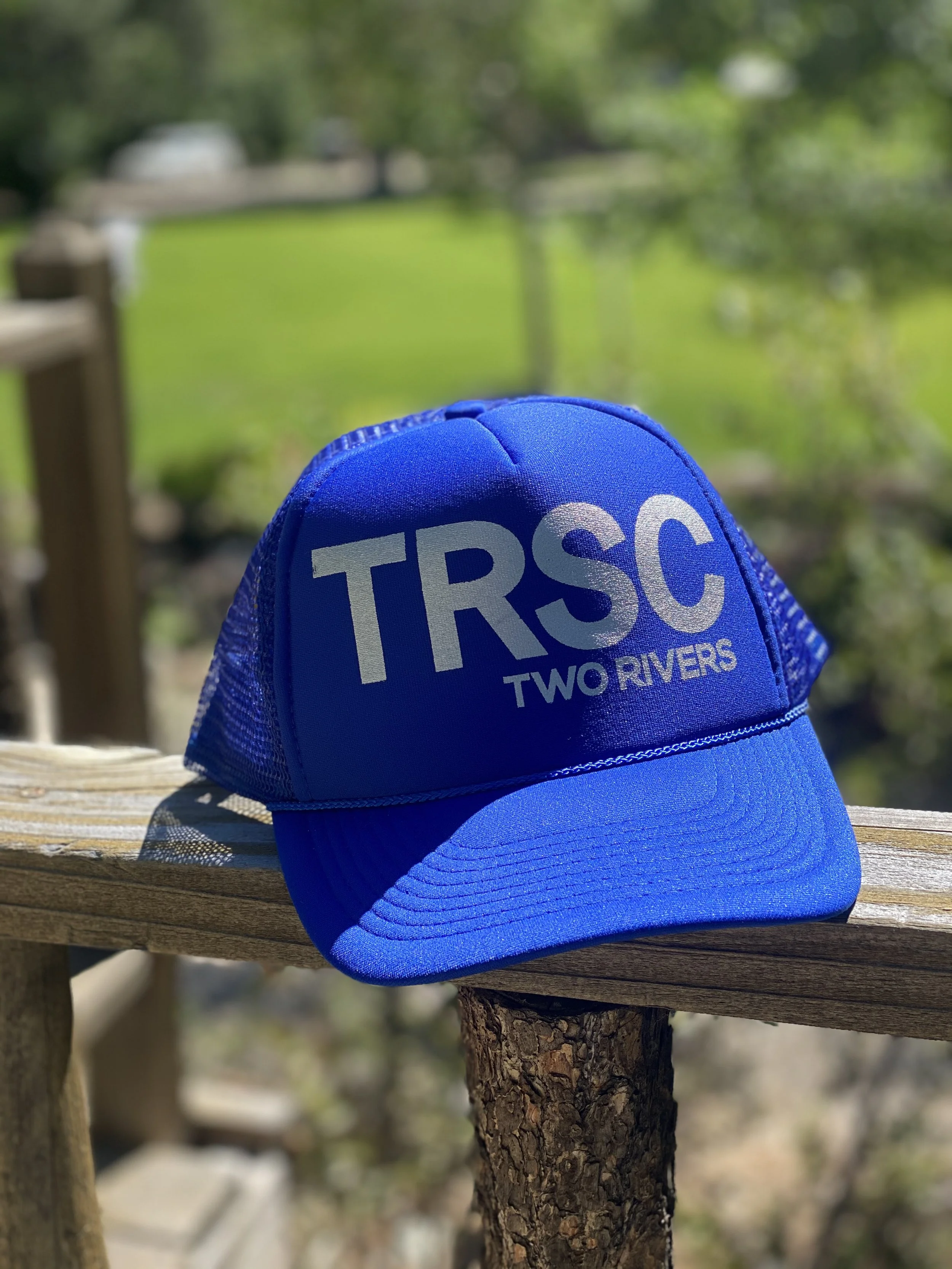 Blue trucker cap with the text "TRSC" and "TWO RIVERS" sitting on a wooden rail outdoors, with a blurred green yard and trees in the background.