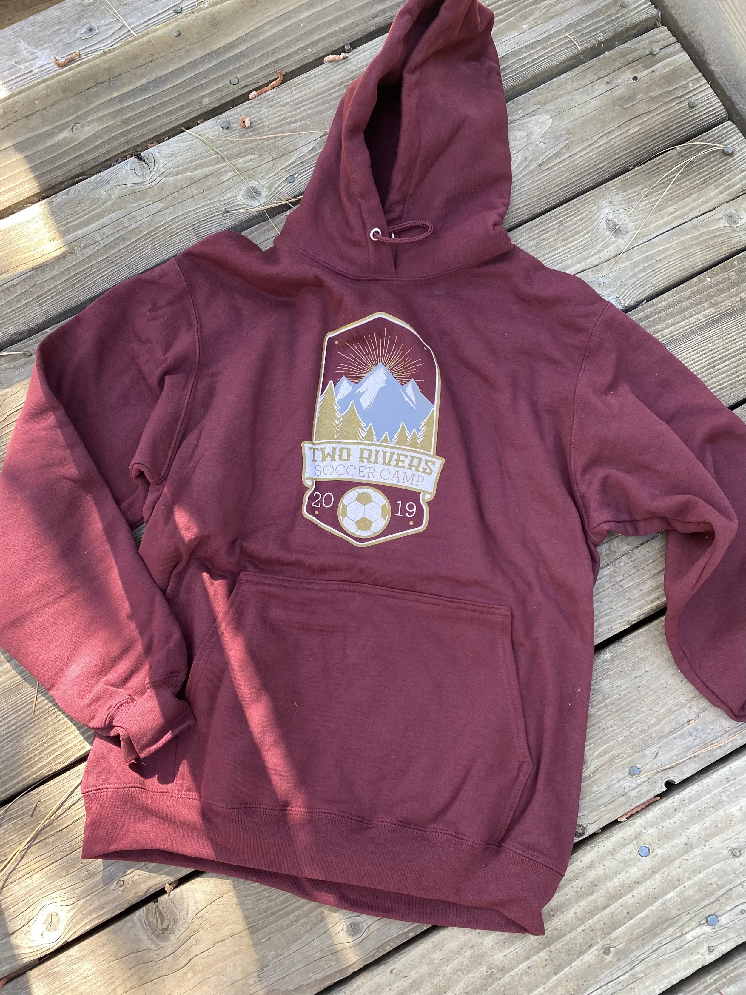 Maroon hoodie with a graphic design featuring mountains, trees, sun rays, and a soccer ball, with the text 'Two Rivers Soccer Camp 2019,' lying on a wooden deck.