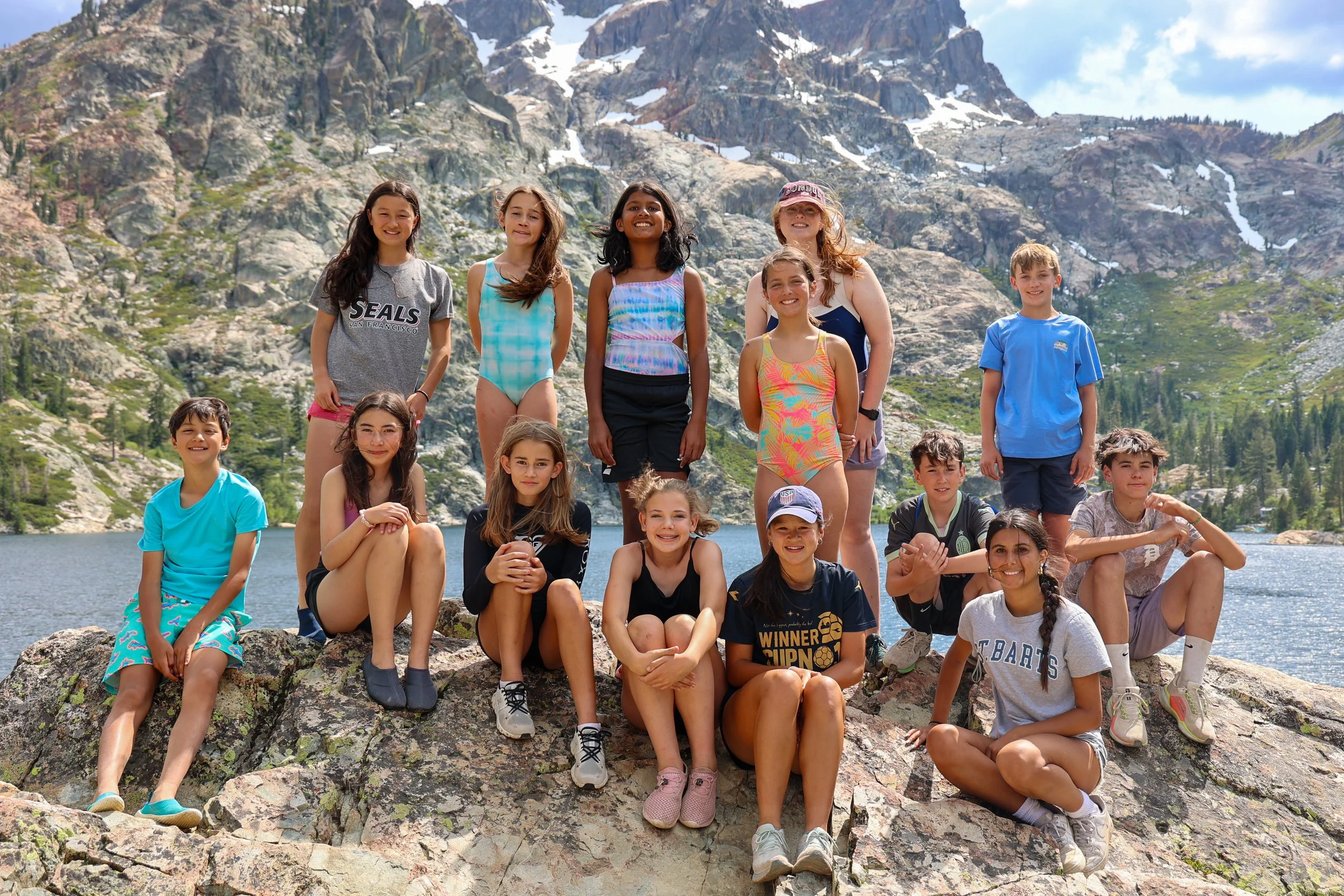 Group of children and a woman posing on rocks near a lake with mountains in the background on a sunny day.