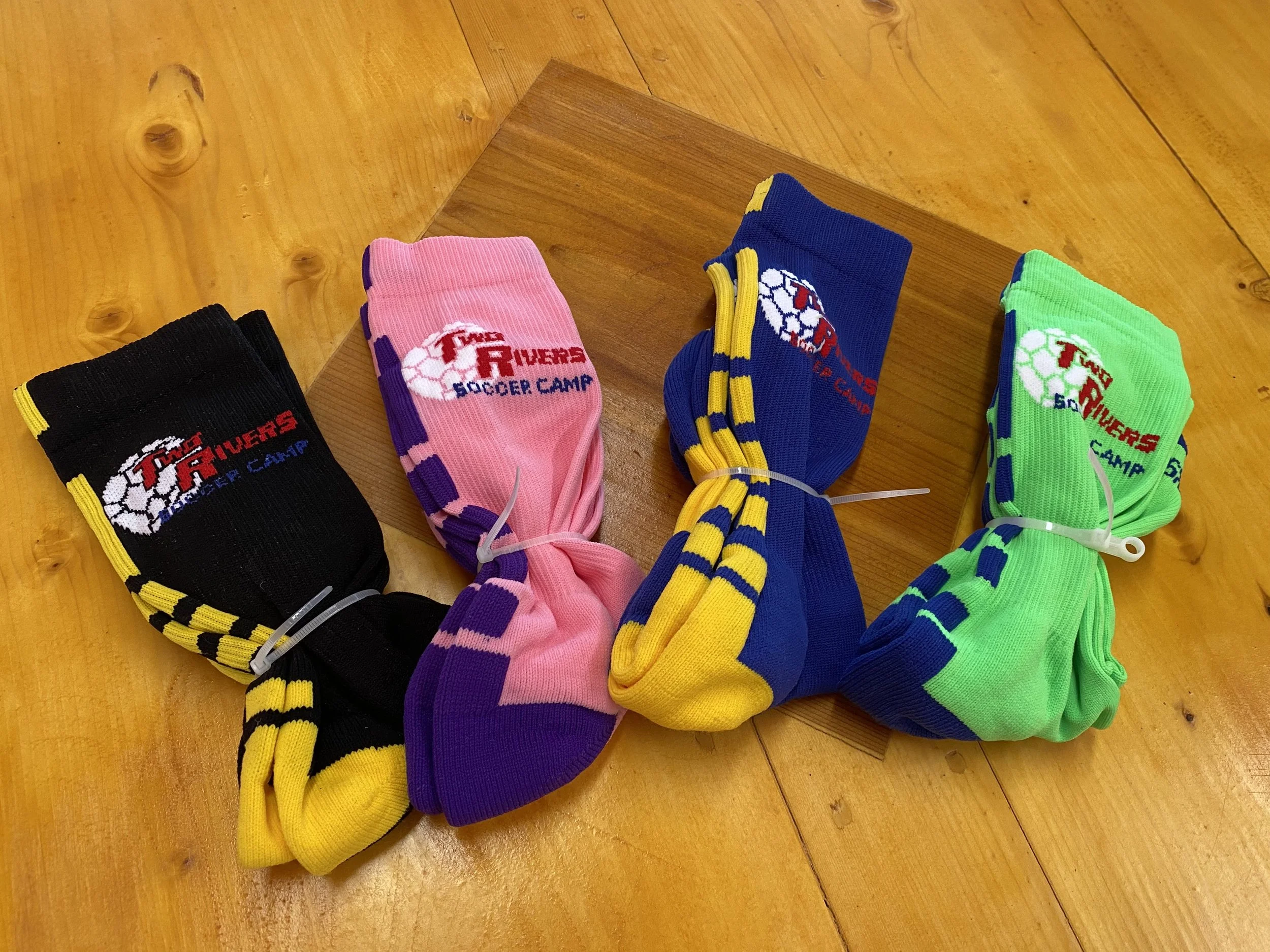 Four pairs of colorful socks with soccer-themed design, labeled 'Fins Rivers Soccer Camp', arranged on a wooden floor.