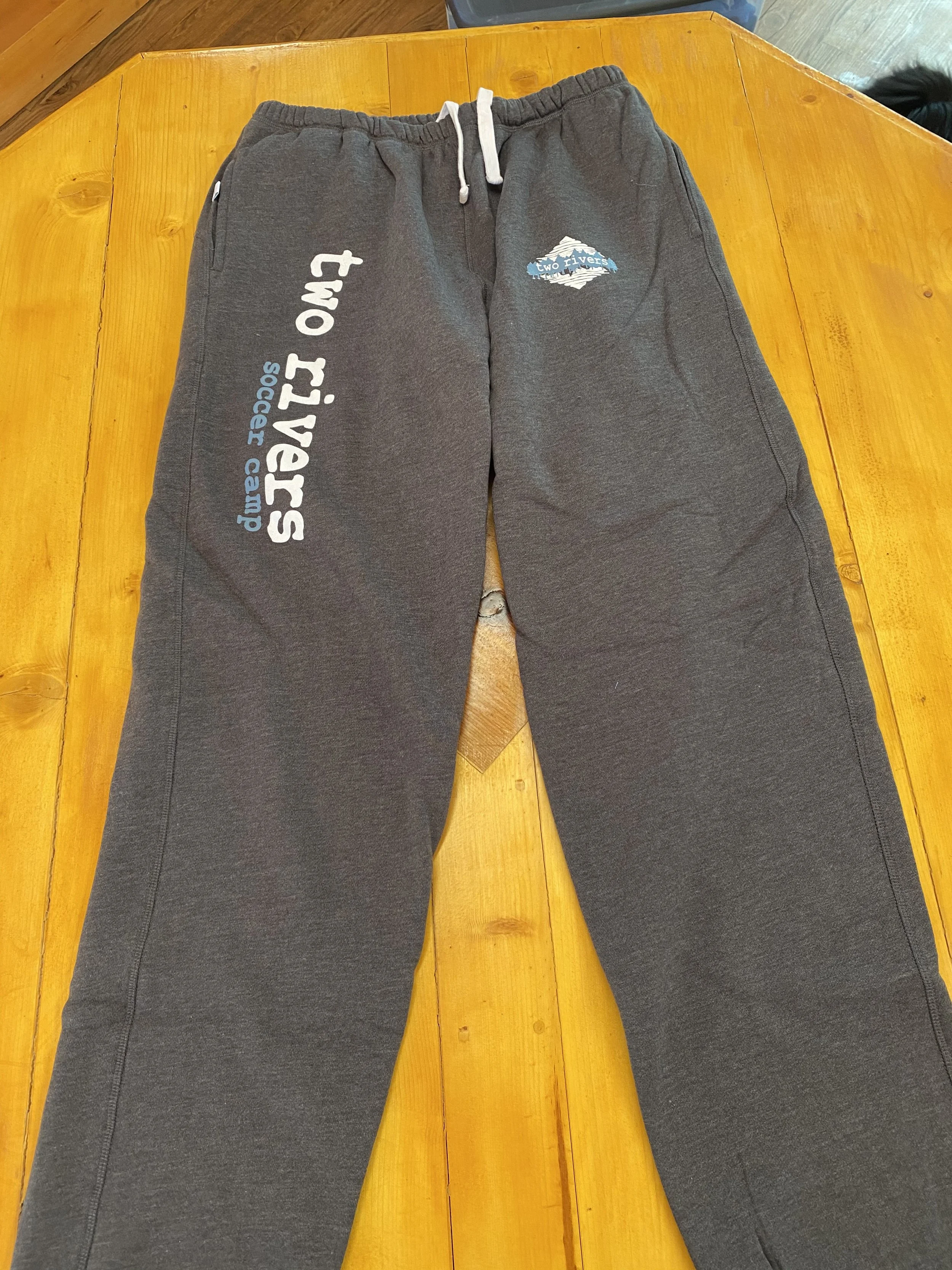 Gray sweatpants with drawstrings on a wooden table, featuring white and blue text and logos related to Two Rivers Social Camp.