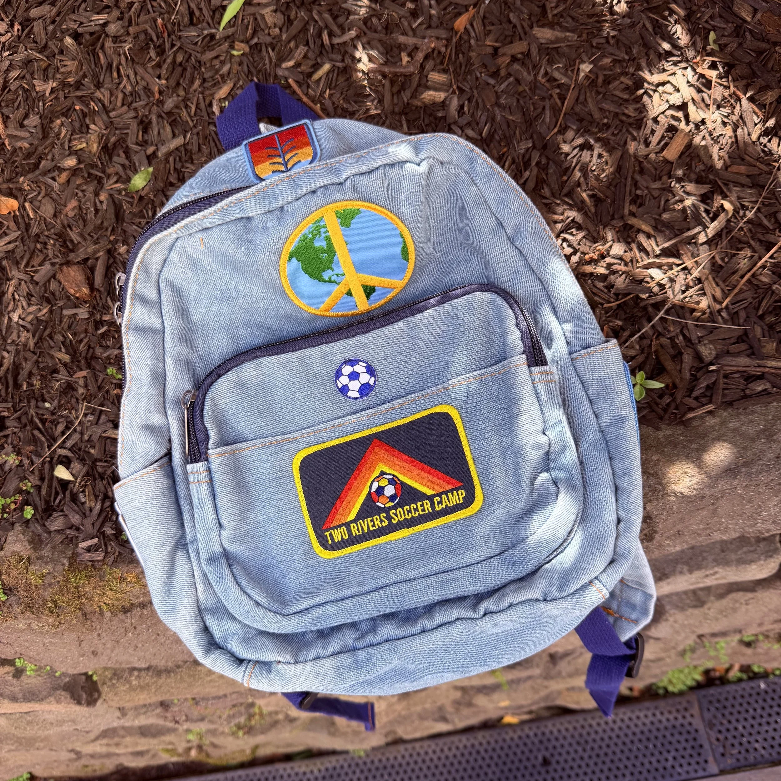 A grey backpack with patches, one of which reads 'Two Rivers Soccer Camp' and another features a soccer ball. The backpack is on a sidewalk next to mulch and a stone wall.