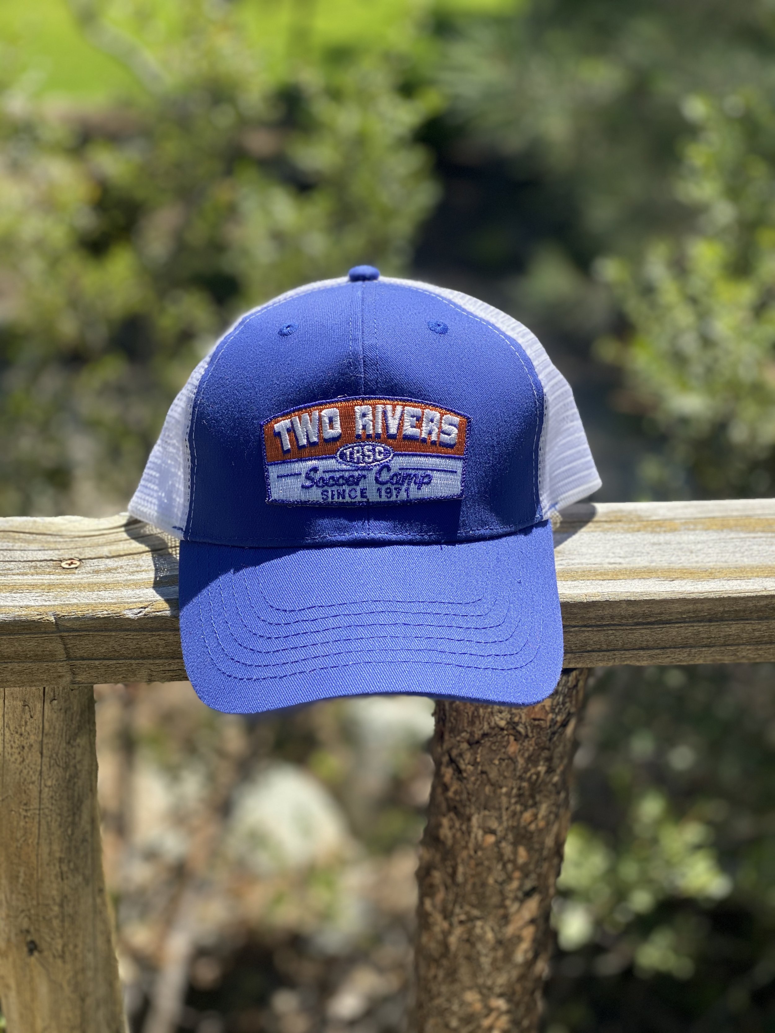 A blue and white trucker cap with an embroidered patch reading "Two Rivers Soccer Camp Since 1971" is placed on a wooden railing outdoors with a blurred natural background.