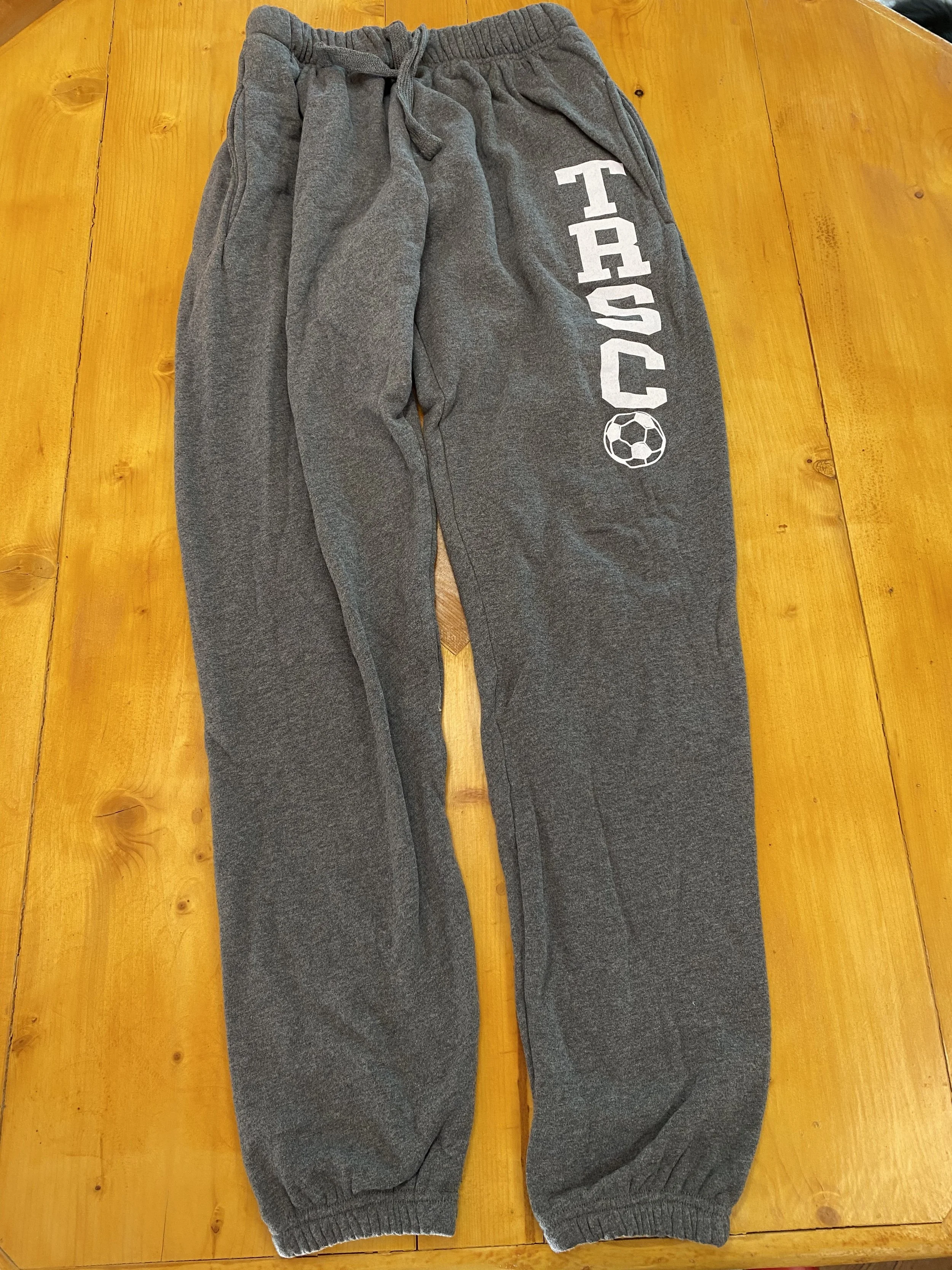 Pair of gray sweatpants on a wooden surface, with the word 'TACOS' and a soccer ball graphic printed on the right leg.