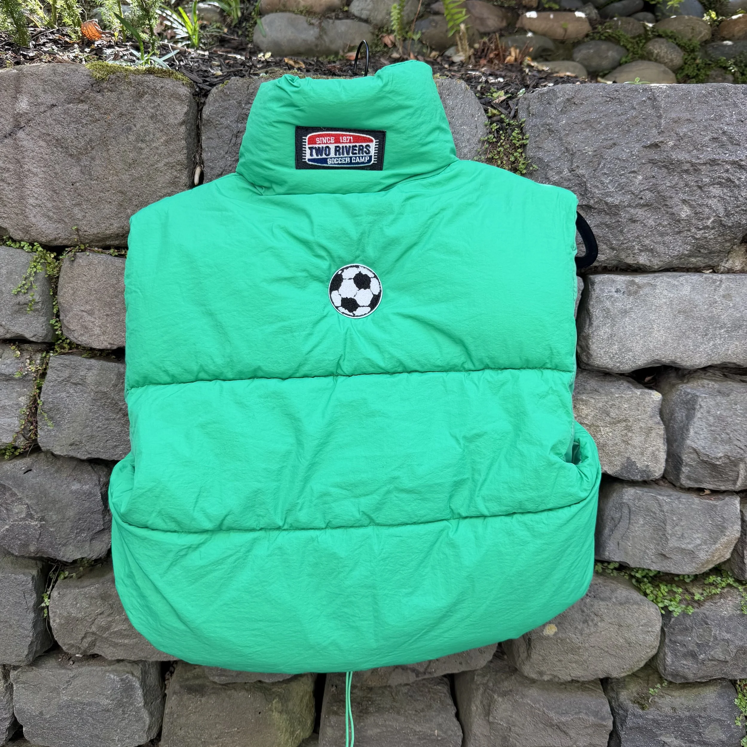 A green puffer vest with a soccer ball patch and a tag that reads 'Two Rivers Soccer Camp' is hanging against a stone wall.