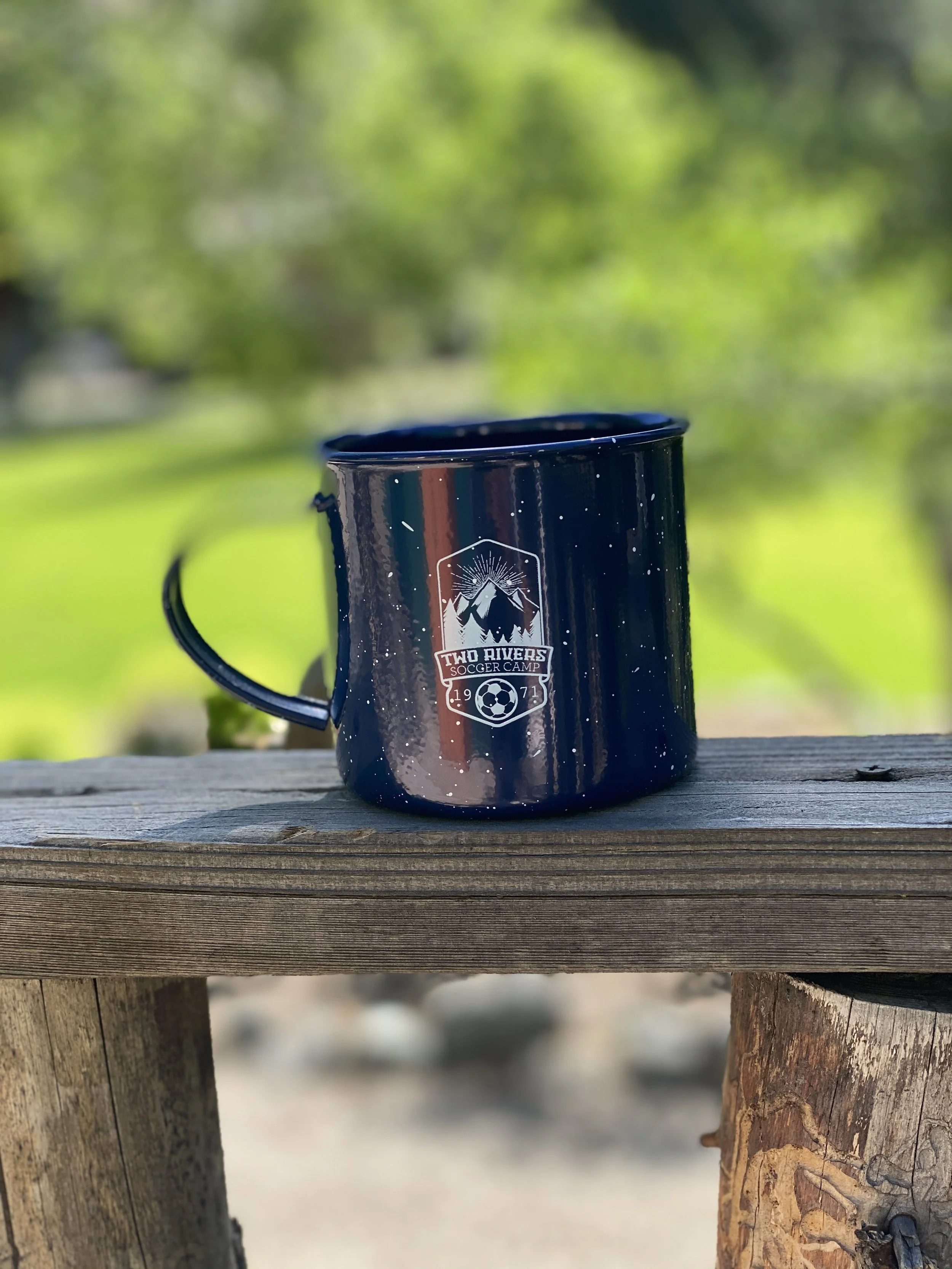 Black enamel camping mug with a Two Rivers Soccer Camp logo, sitting on a wooden surface outdoors with green trees in the background.