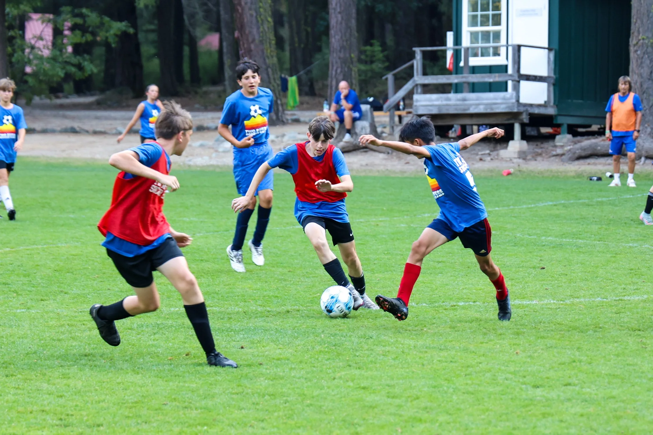 Young boys playing soccer on a grassy field, with trees and a small building in the background.
