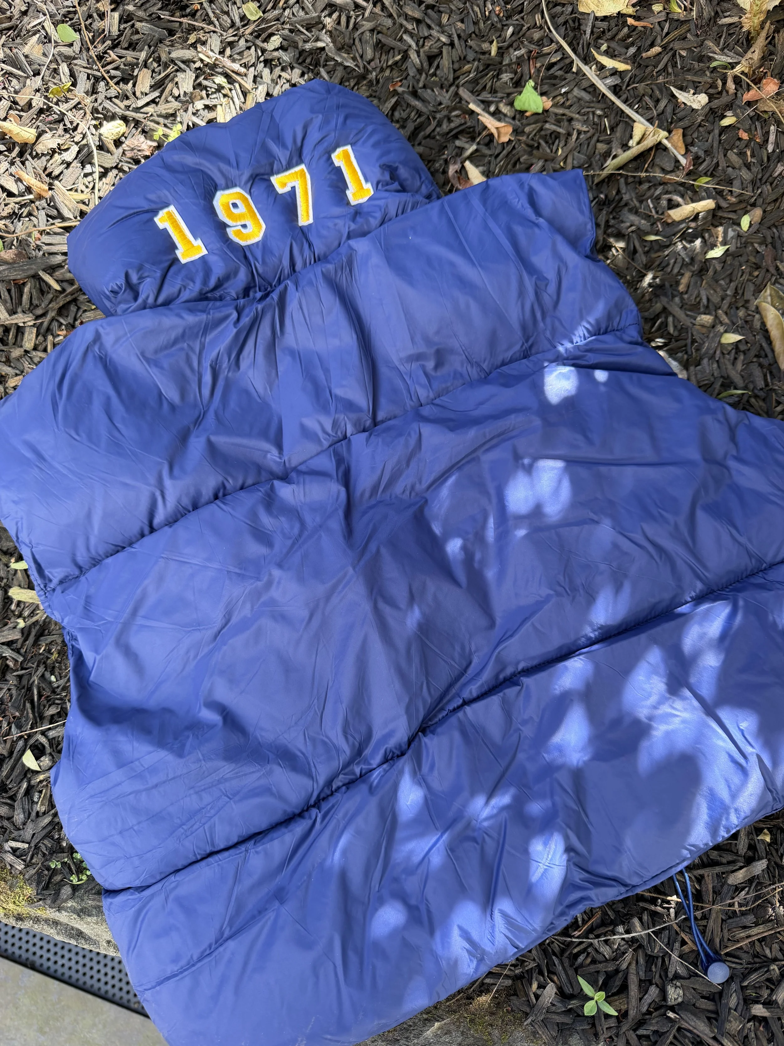 A blue puffer vest with yellow and white embroidered numbers '1971' on a bed of brown mulch with scattered leaves and small plants.