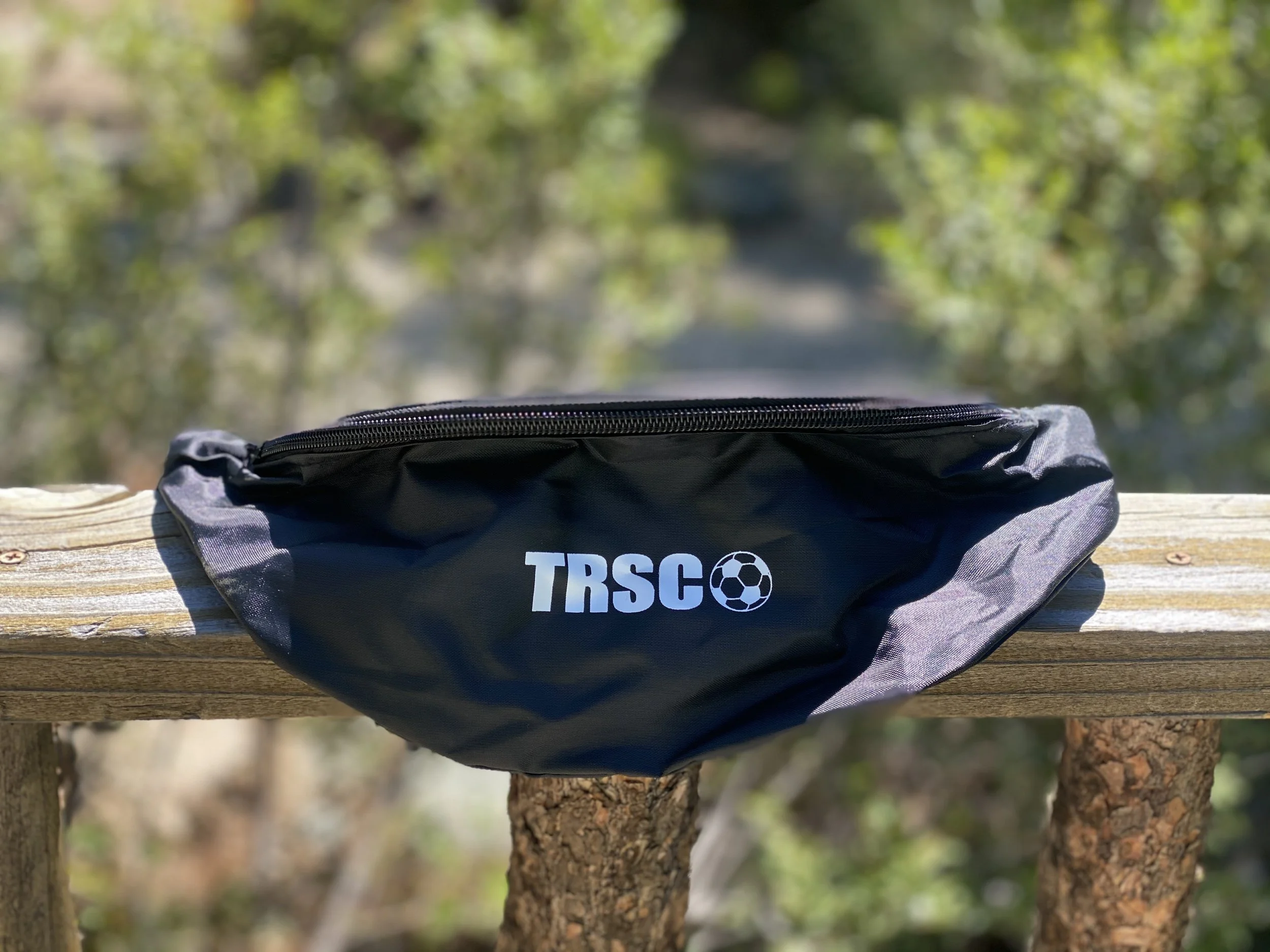 A black and gray fanny pack with a zipper and the logo 'TRSCO' and a soccer ball graphic, placed on a wooden railing outdoors with blurred green foliage in the background.