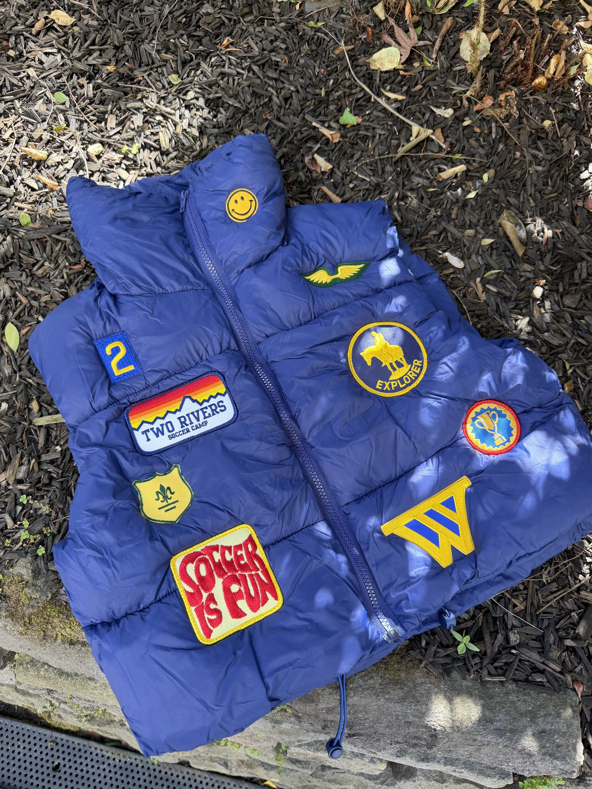 A blue puffer vest with various colorful patches lies on the ground among mulch and small leaves.