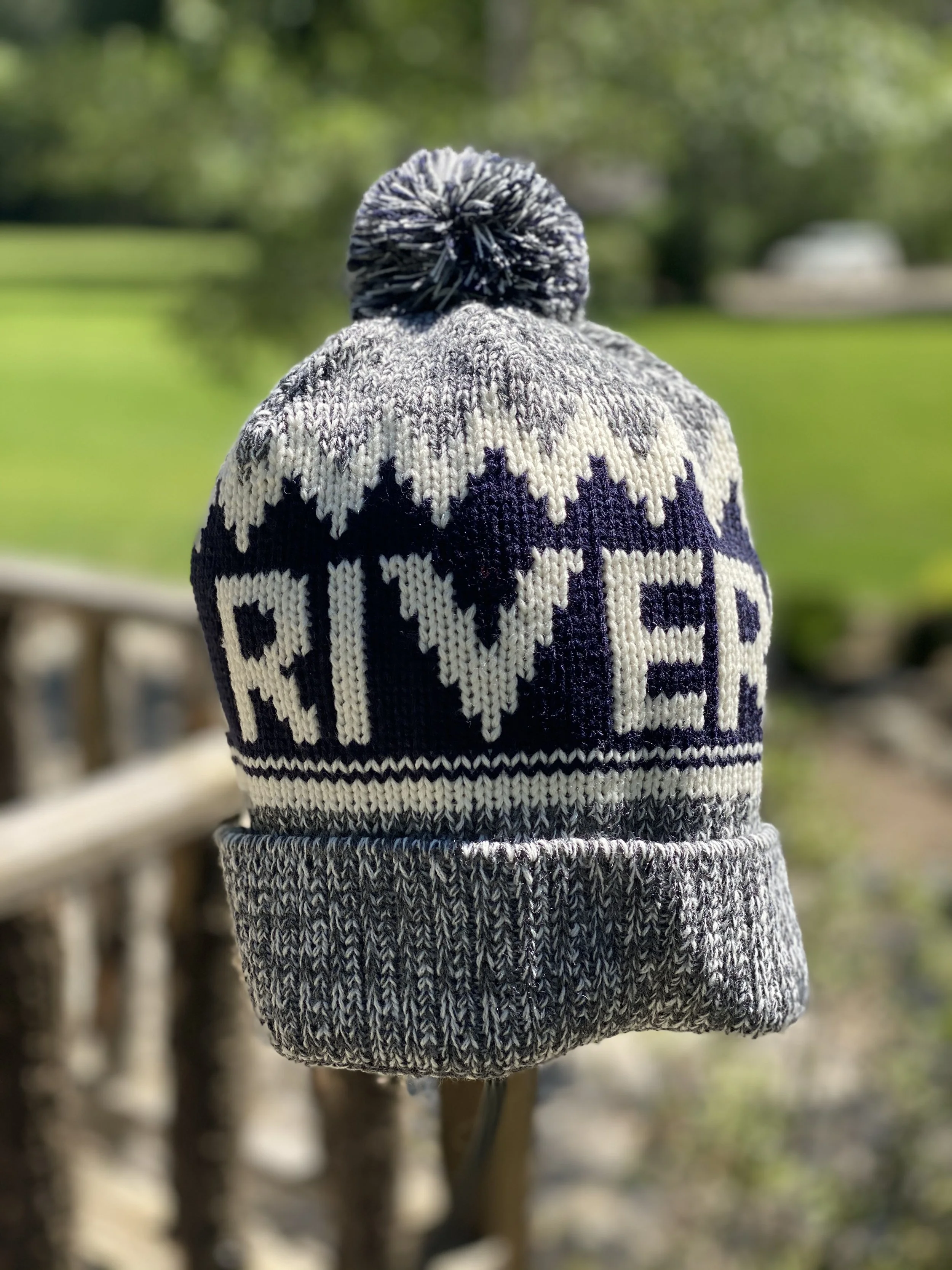 A knit beanie hat with a pom-pom on top, featuring a pattern that spells out 'RIVER' in bold letters, outdoors with green grass and trees in the background.
