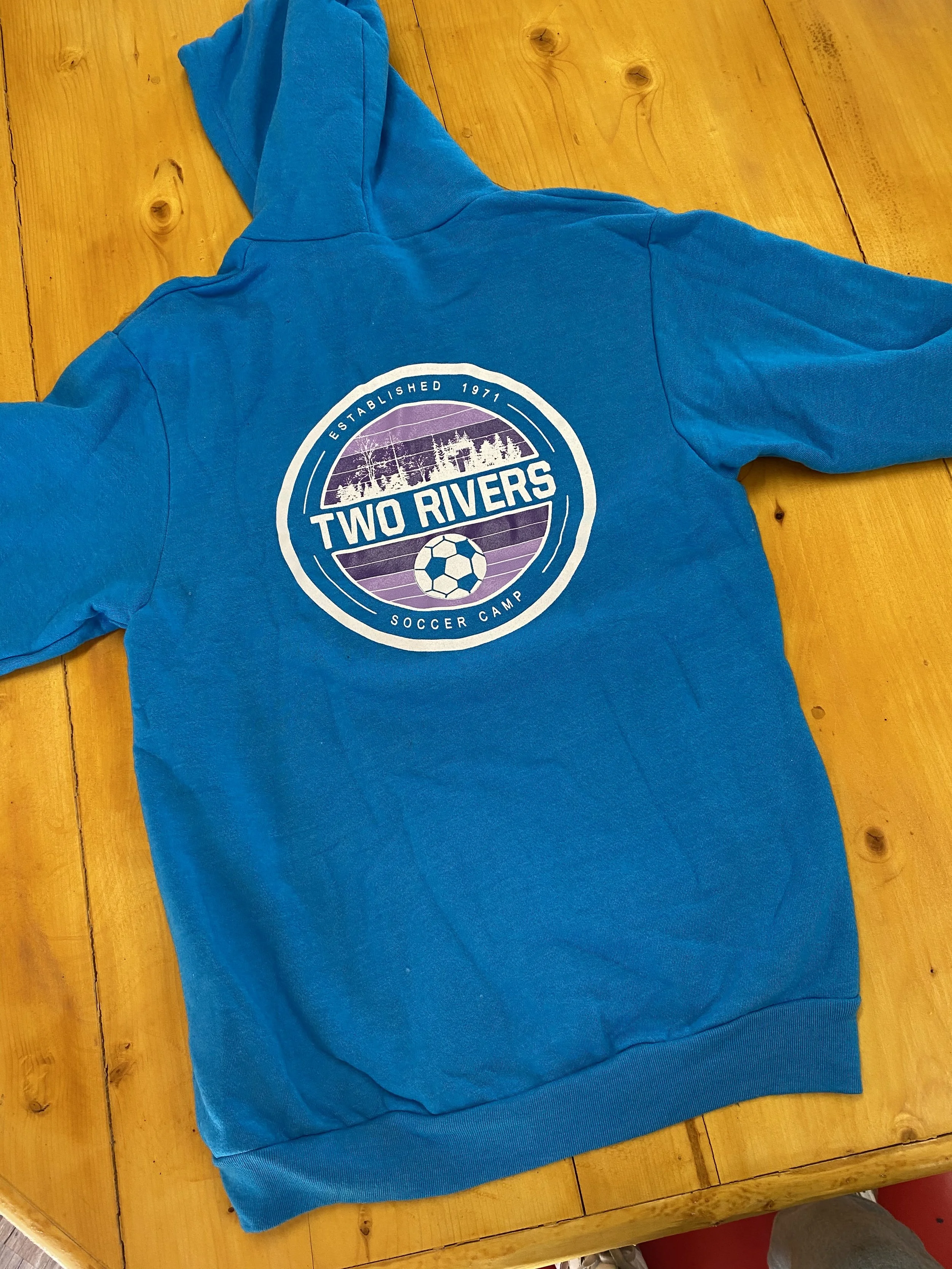 Blue hoodie with a circular logo on the back for Two Rivers Soccer Camp, established in 1971, with a soccer ball graphic and a flooded riverscape in purple and white, laid on a wooden table.